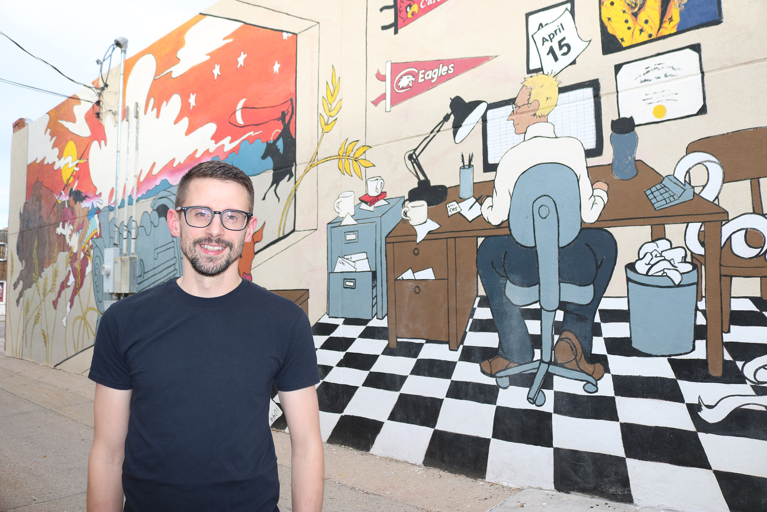 Travis Hencey stands in front of “Beating the Deadline,” a mural he designed for Chadron’s Art Alley. The mural features a man at a desk working on a computer.