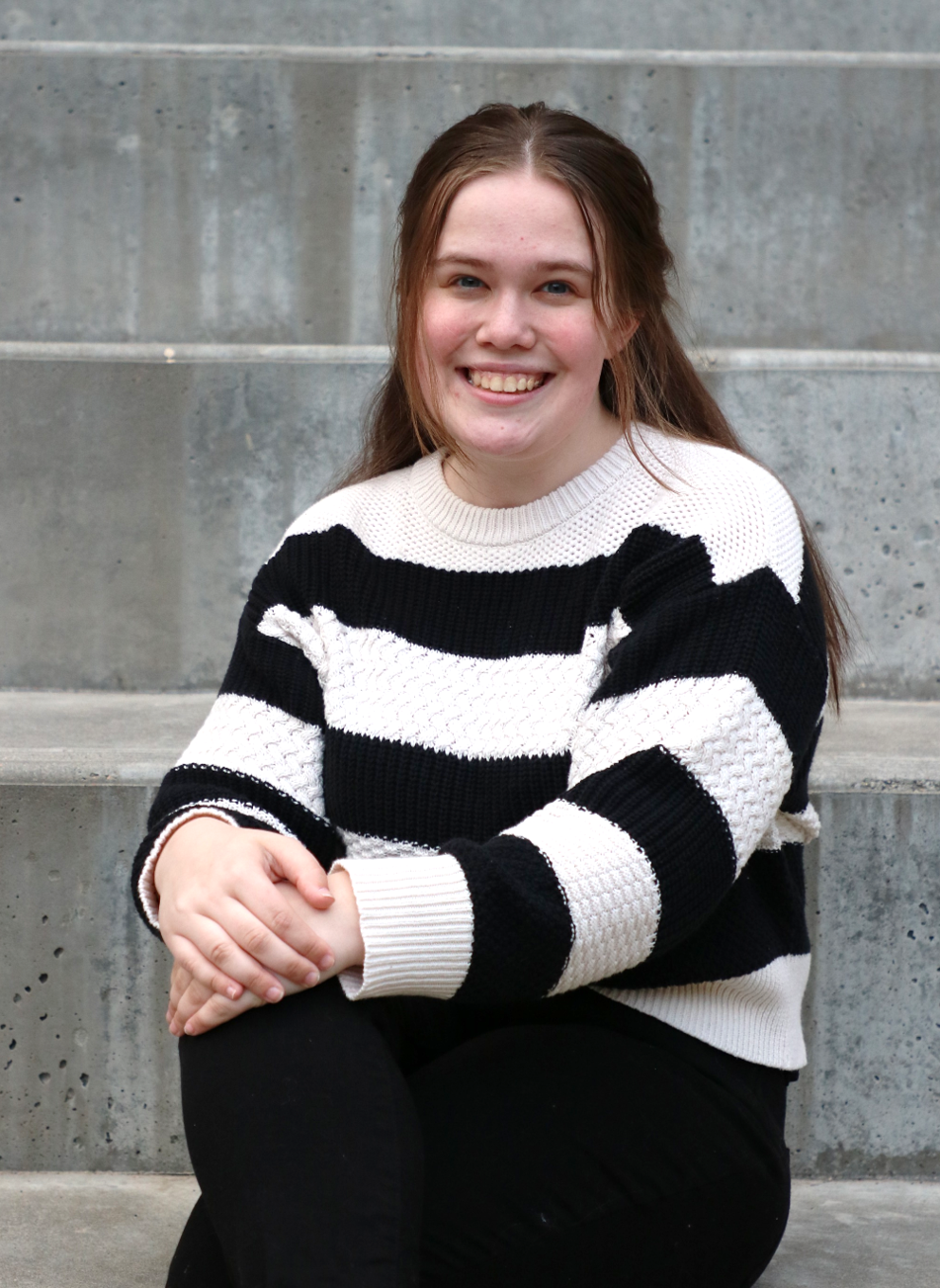 Color portrait of Hannah Scholen, University Honors Program member and May graduate, wearing a black-and-white-striped sweater and black pants, sitting on concrete steps.