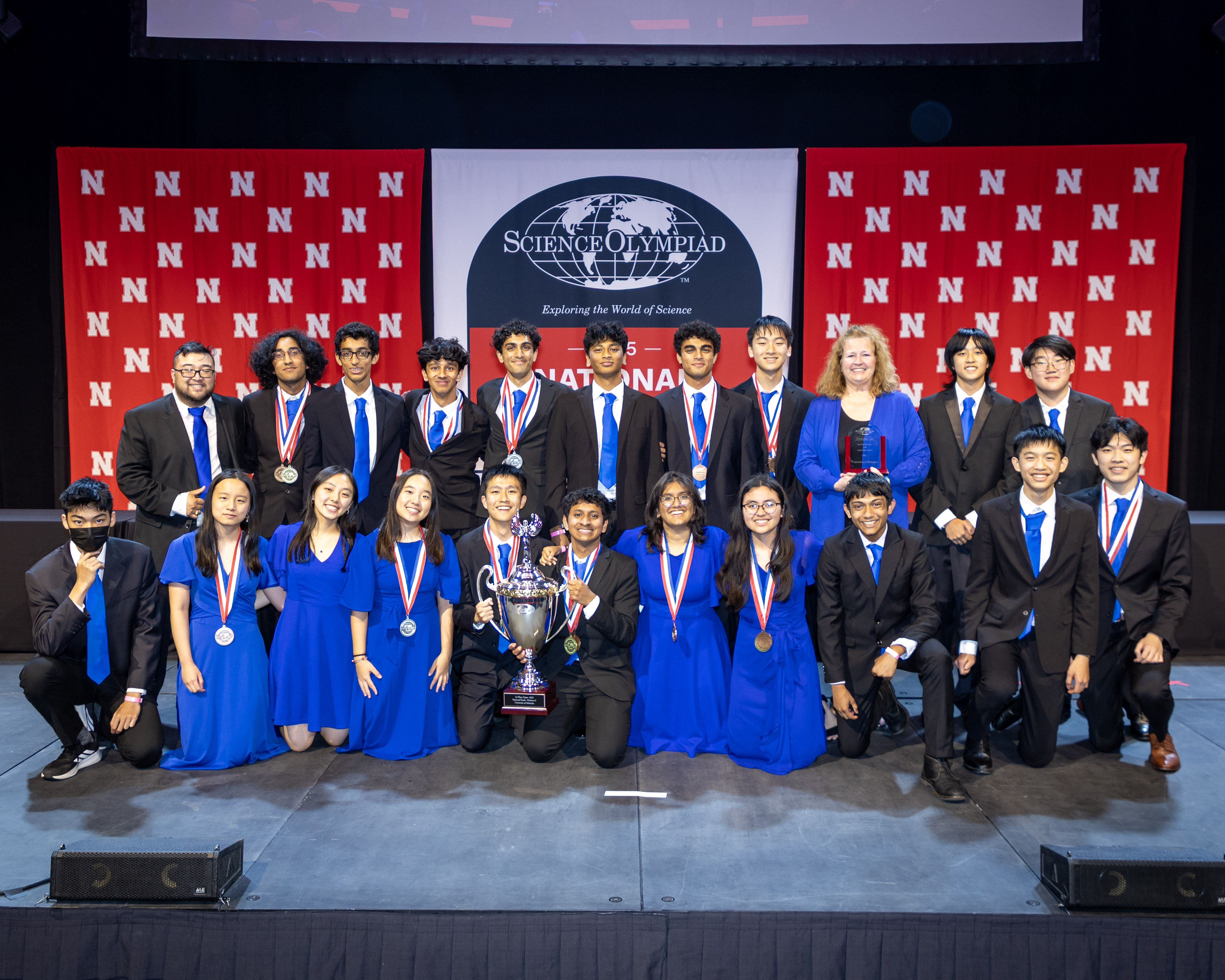 Twenty students and their coaches from Seven Lakes High School in Katy, Texas, wearing formal attire, pose with a large trophy in front of a Husker background and the Science Olympiad National Tournament logo.
