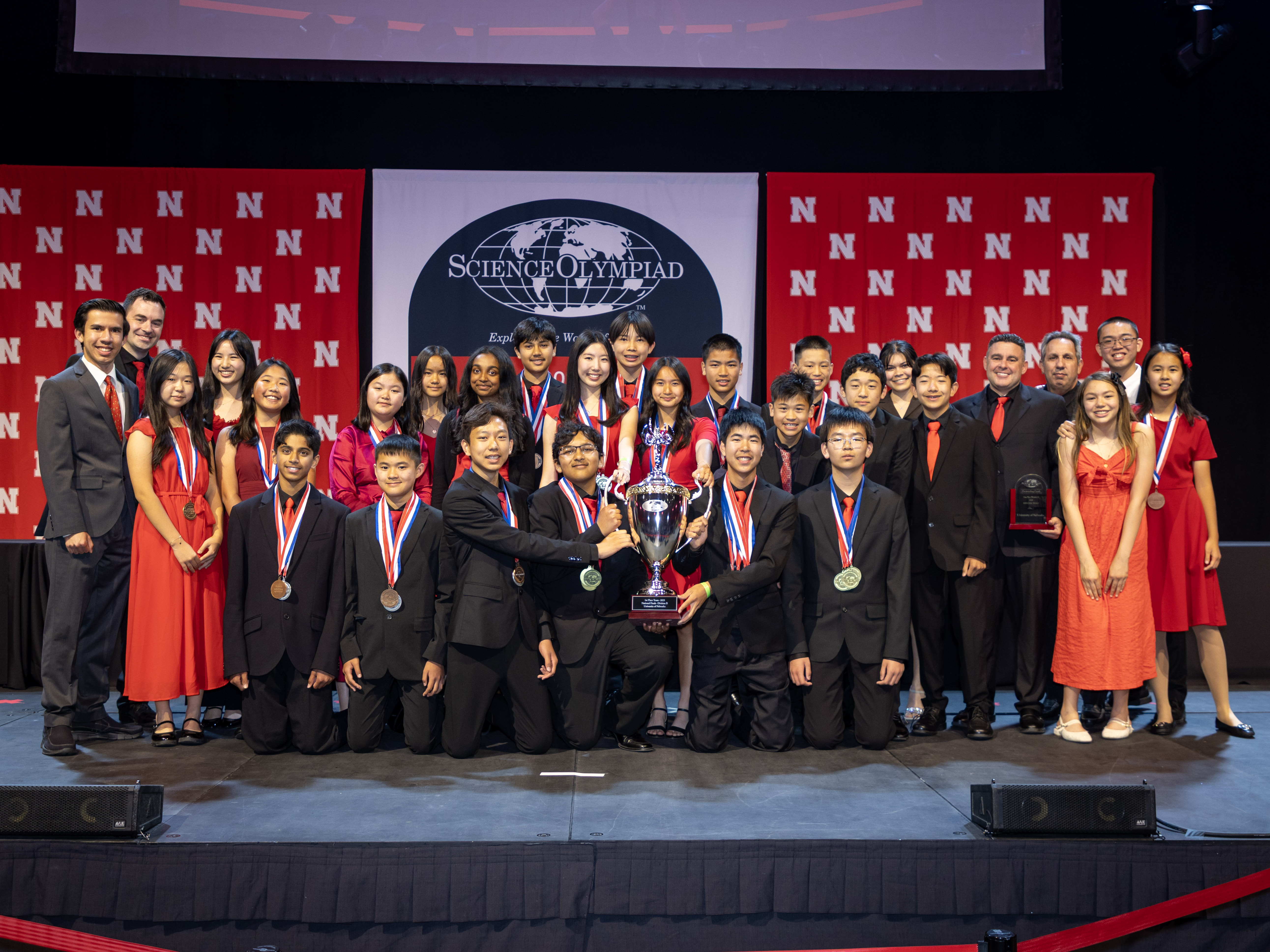 About two dozen students and their coaches from Sierra Vista Middle School in Irvine, California, wearing formal attire, pose with a trophy in front of a Husker background and the Science Olympiad National Tournament logo.