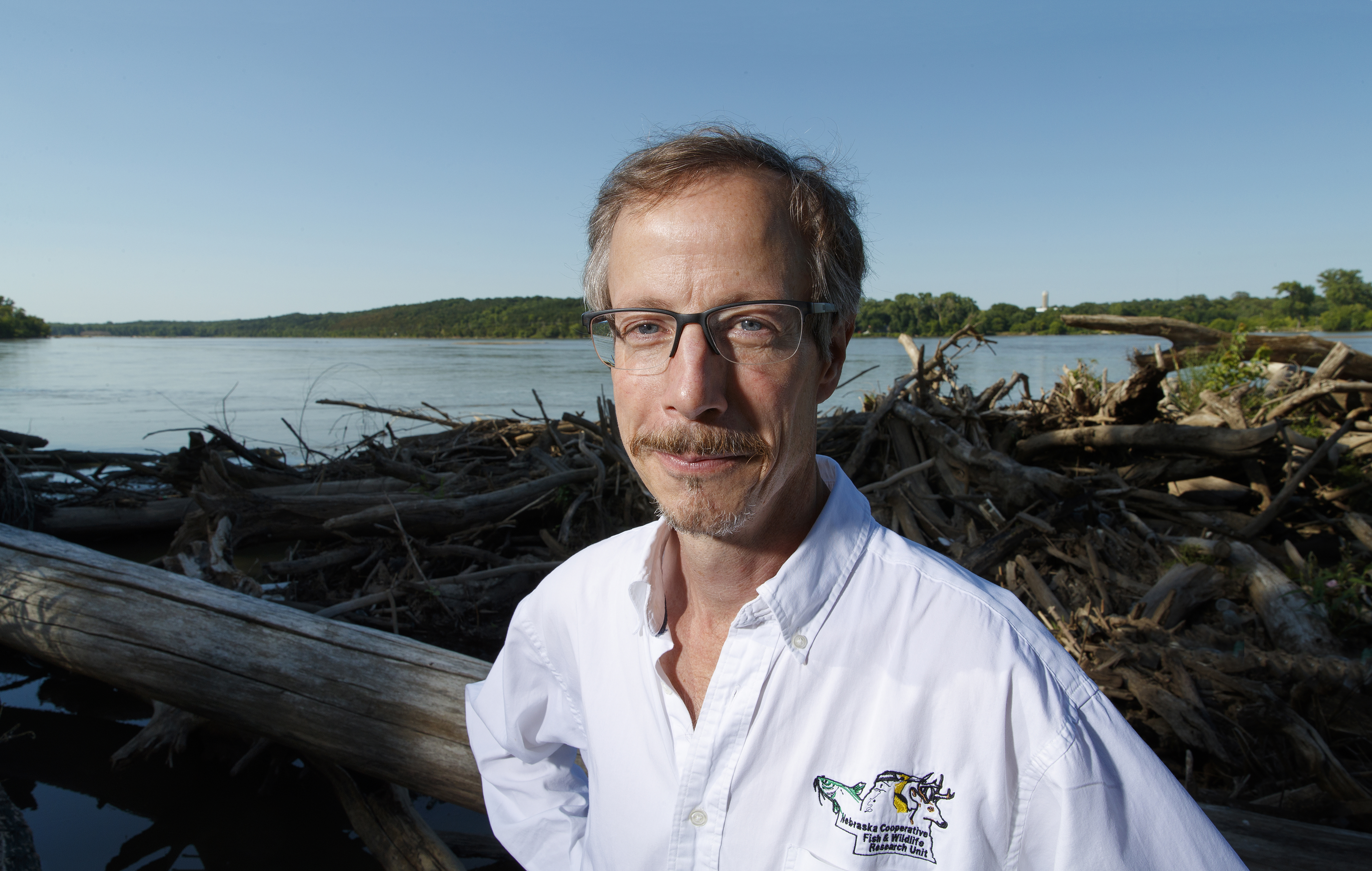Craig Allen, professor in the School of Natural Resources, poses near logs next to a river. He wears a white button-up shirt with a logo featuring a fish, bird and deer and the words "Nebraska Cooperative Fish and Wildlife Research Unit."