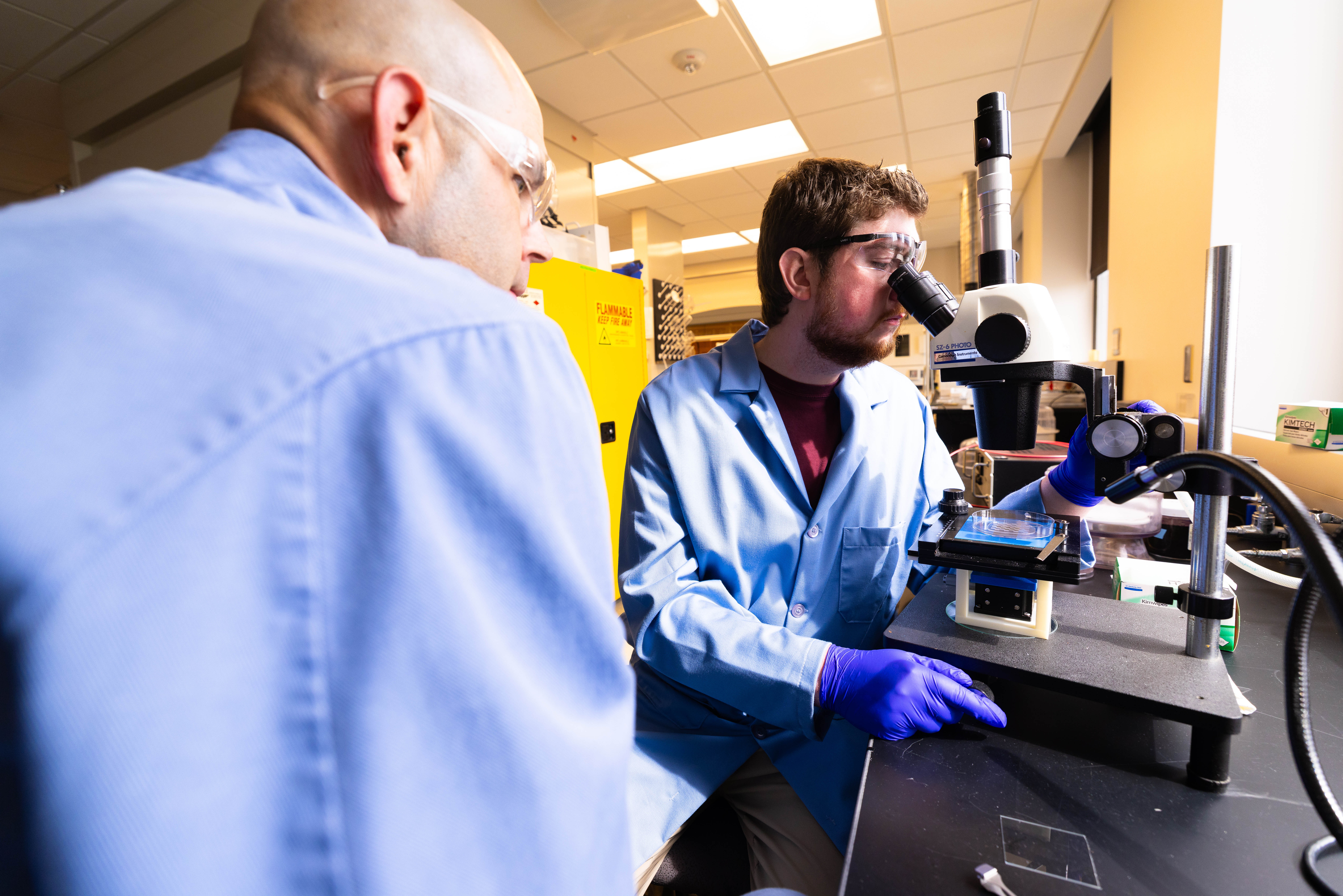 Brennan Watts, a fourth-year doctoral student, looks through a microscope at a checkerboard-patterned hydrogel, with Stephen Morin, associate professor of chemistry.