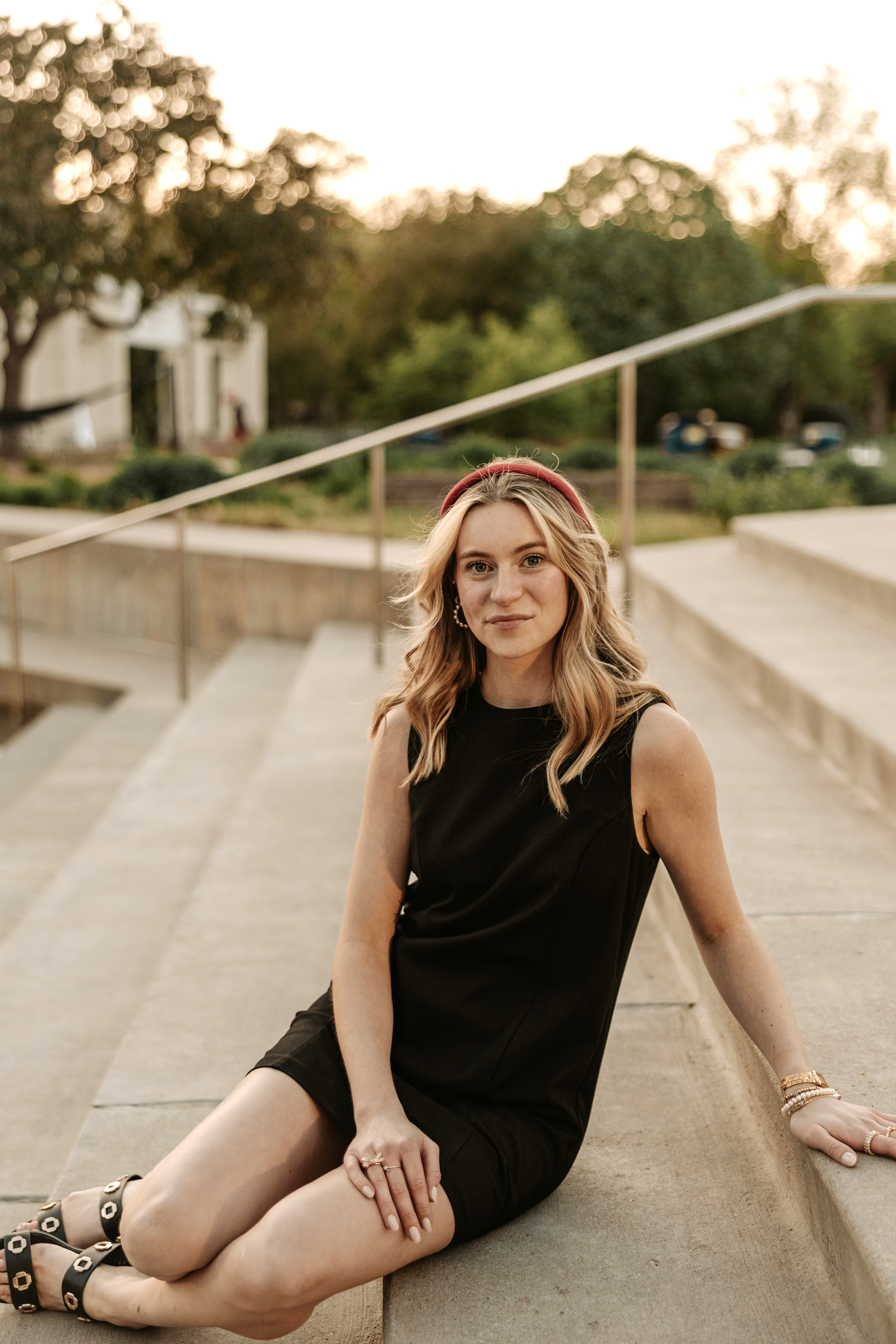Brigid Toomey sits on an outdoor staircase. She is wearing a black dress, red headband and strappy sandals.