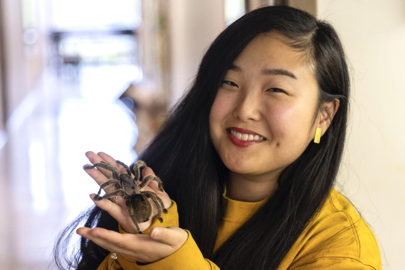 Jillian Kurovski smiles while holding a tarantula in a hallway. She is wearing a yellow shirt.