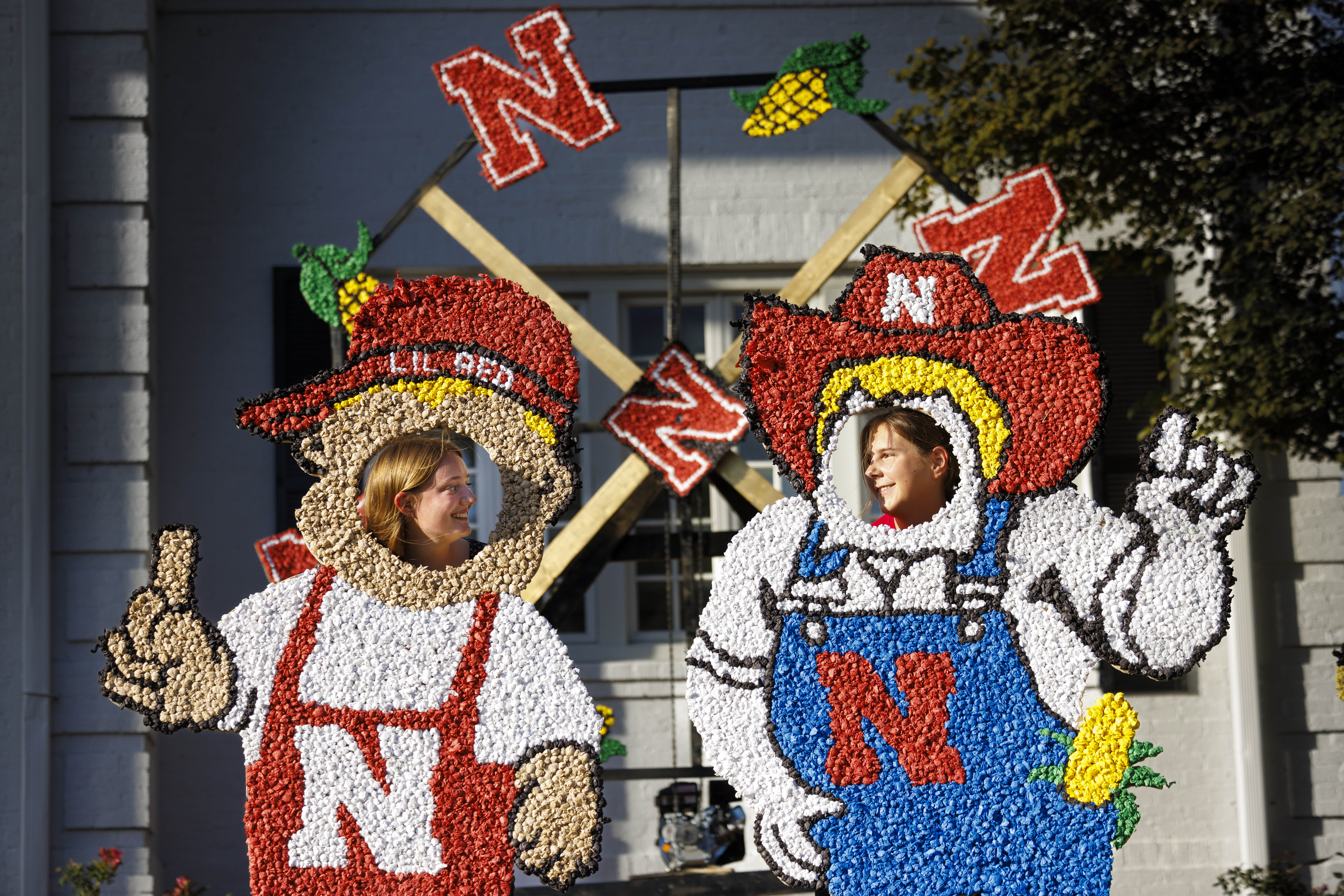 Two female students put their heads through Lil Red and Herbie cutouts decorated with pomping in front of a fraternity house.