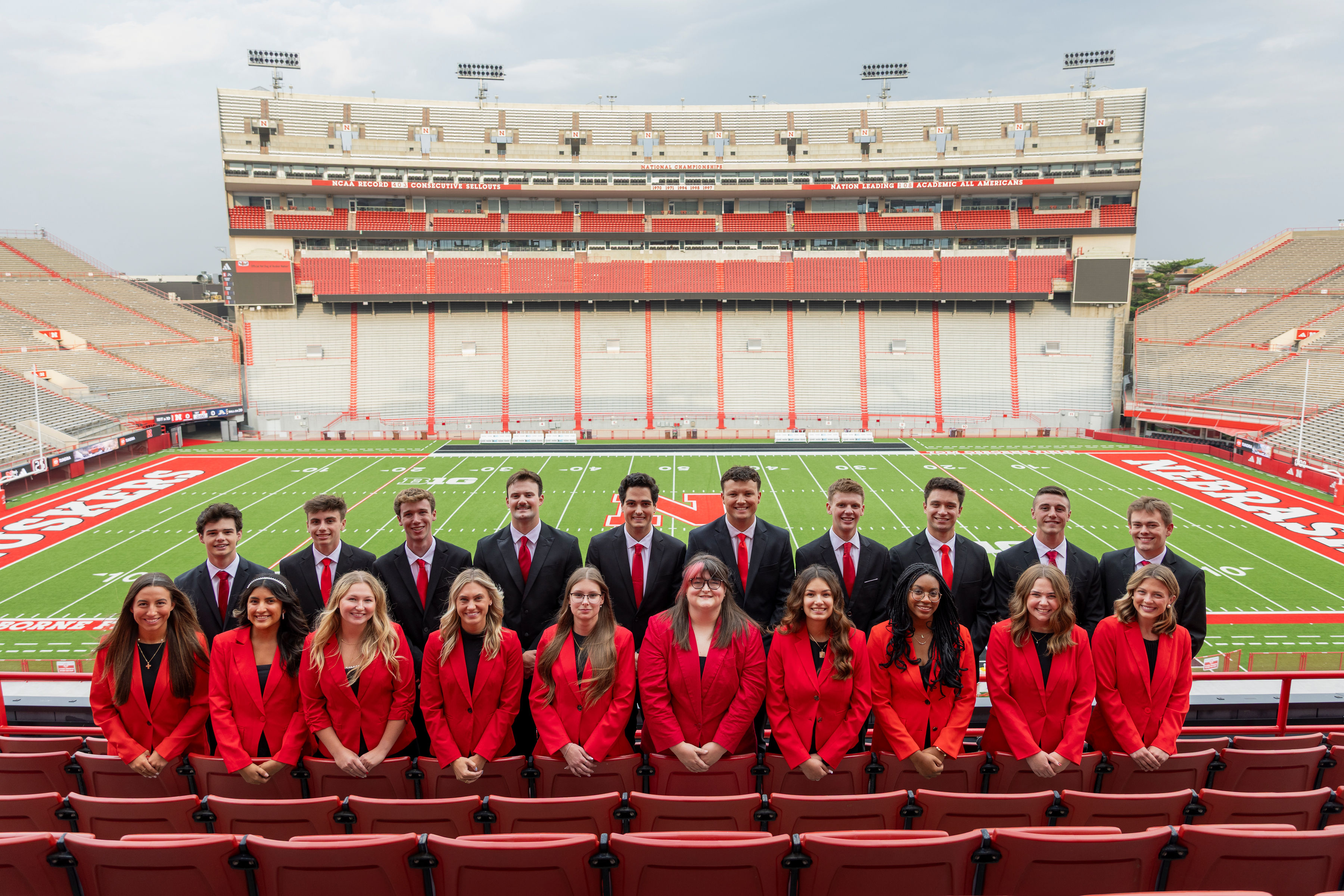 Ten young men and 10 young women — all 2025 homecoming royalty finalists — pose for a photo in the stands at Memorial Stadium, with the football field in the background. The men are wearing black suits with red ties, and the women are wearing red blazers with black shirts.