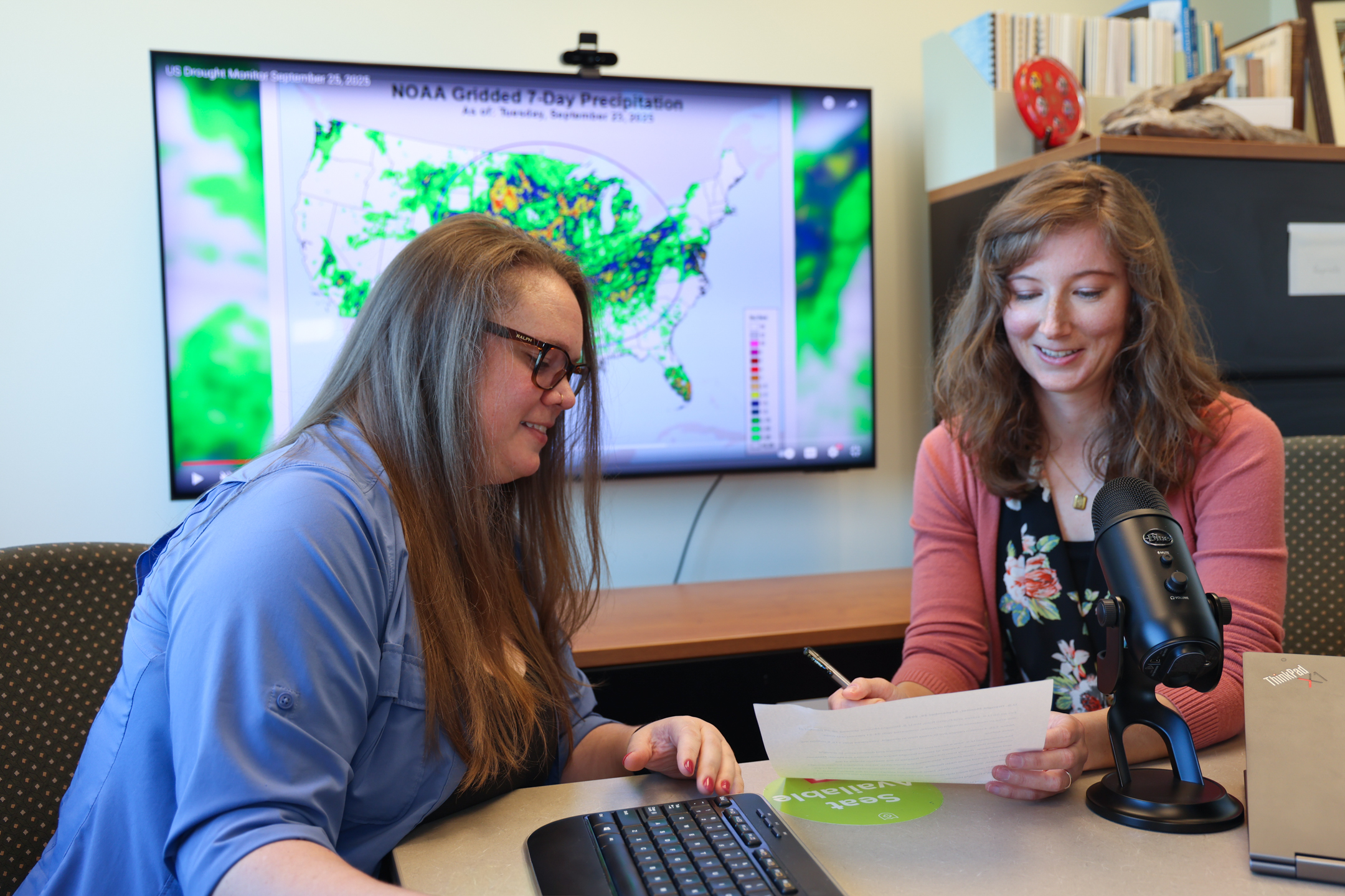 Lindsay Johnson (left) and Emily Case-Buskirk look at a piece of paper at a table with a microphone on it. In the background is a computer monitor with a map of the United States showing "NOAA Gridded 7-Day Precipitation."