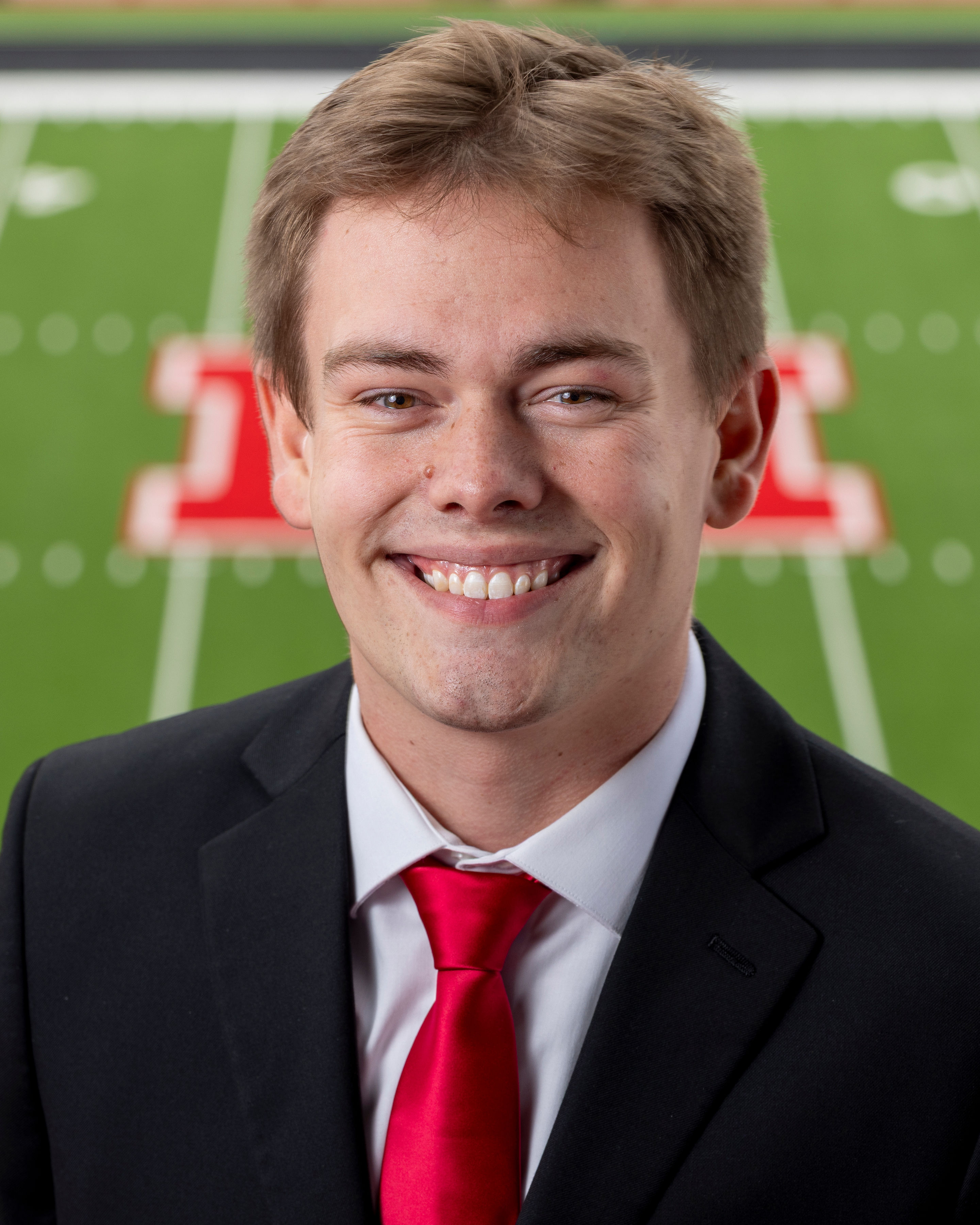 Color portrait of Caden Connelly, wearing a black suitcoat, white shirt and red tie, with Tom Osborne Field in the background.