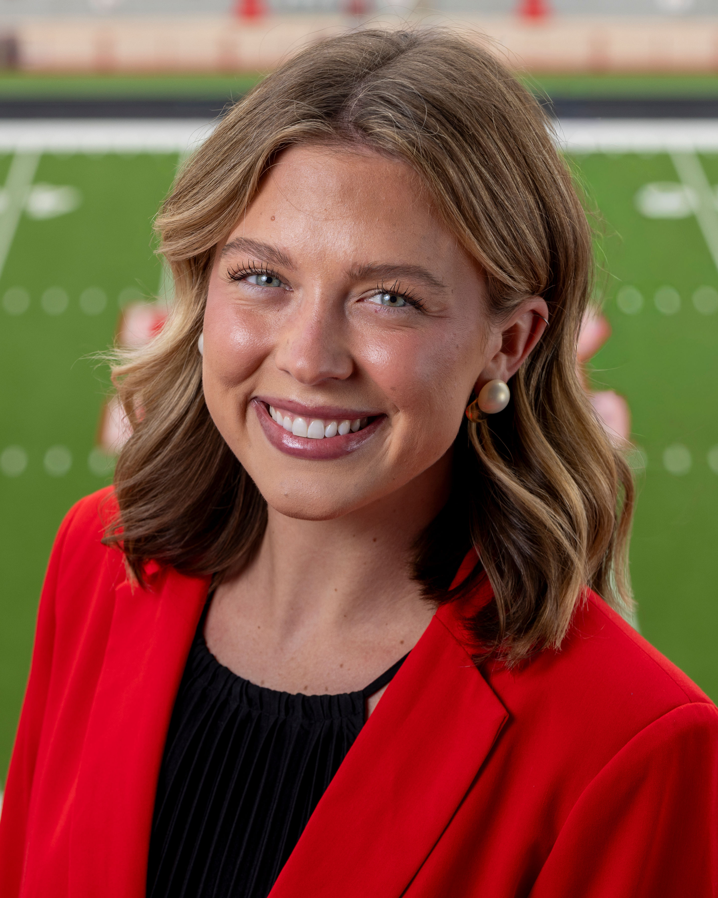 Color portrait of Claire Kelly, wearing a red blazer over a black shirt, with Tom Osborne Field in the background.