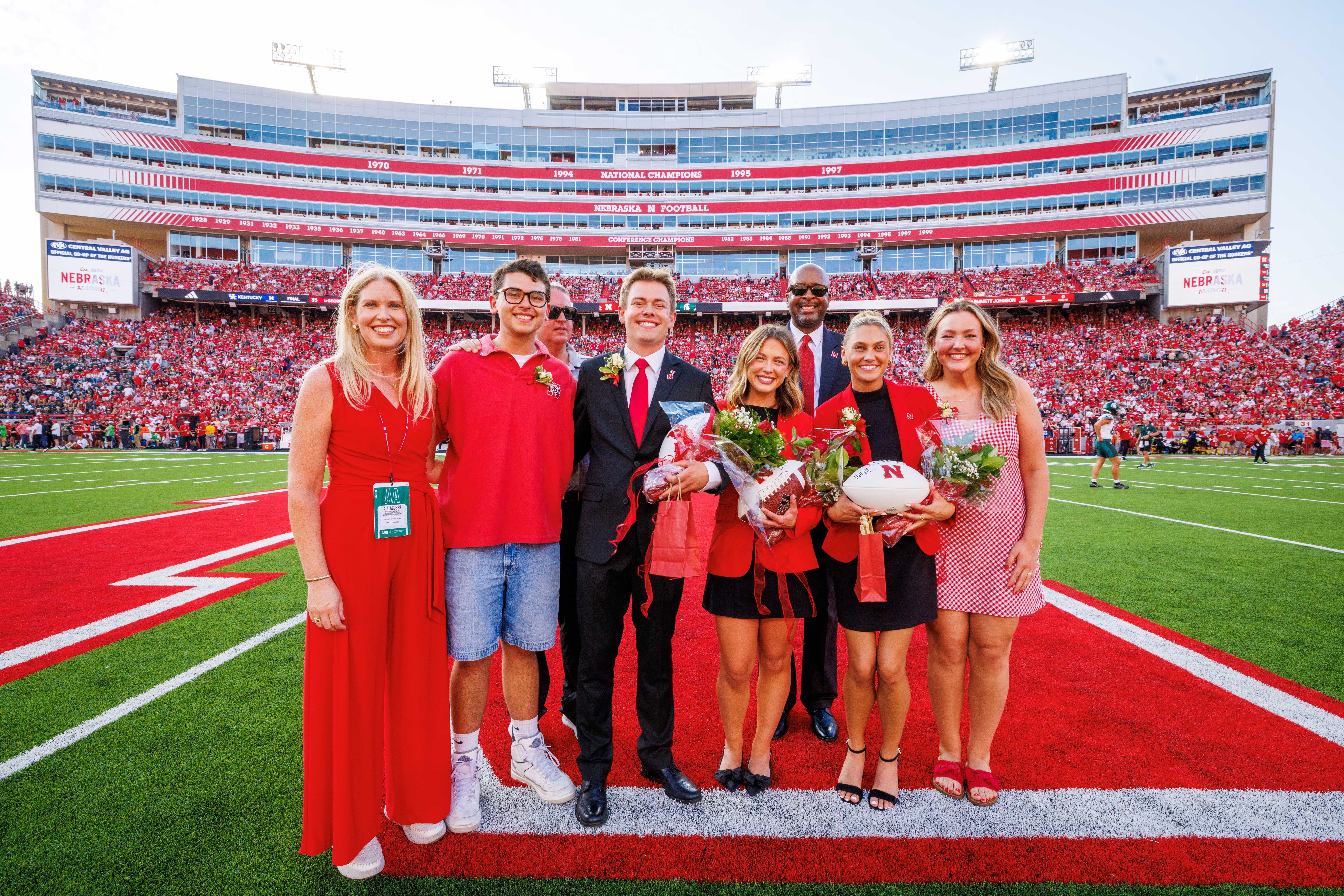 Four women and three men in red and black attire pose for a photograph at midfield in Memorial Stadium.