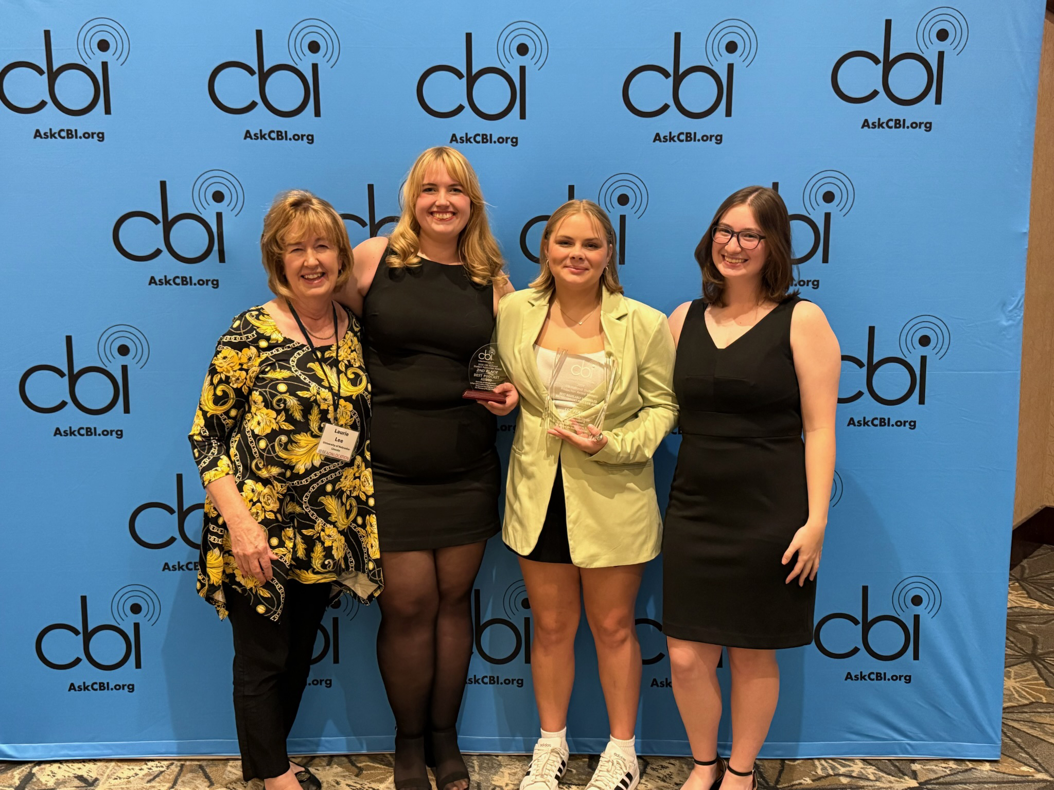 (From left) Nebraska Broadcasting Society faculty adviser Laurie Lee and club members Hayden Hauge, Grace Koch and Katie Mulloy pose with two glass awards in front of a blue background with the words "CBI: AskCBI.org" repeating. They are wearing formal attire.