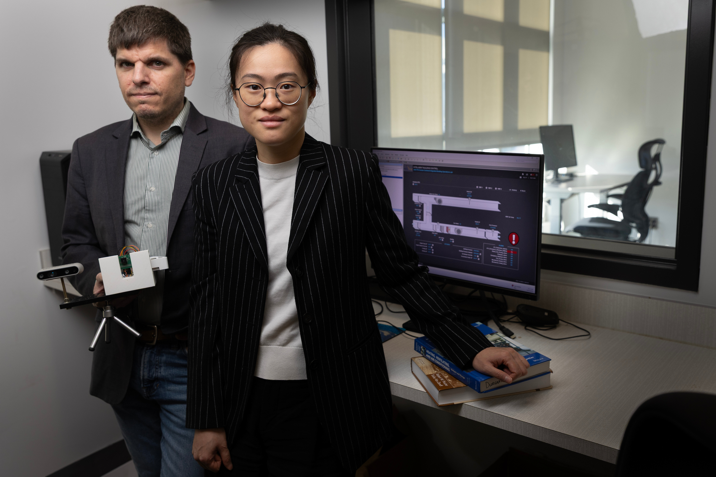 Iason Konstantzos (left) and Xiaoqi (Clare) Liu pose next to a computer and window. Through the window, a desk with a computer on it and an office chair are visible. Konstantzos holds a camera and sensor on a small tripod, and Liu rests her left arm on two textbooks.