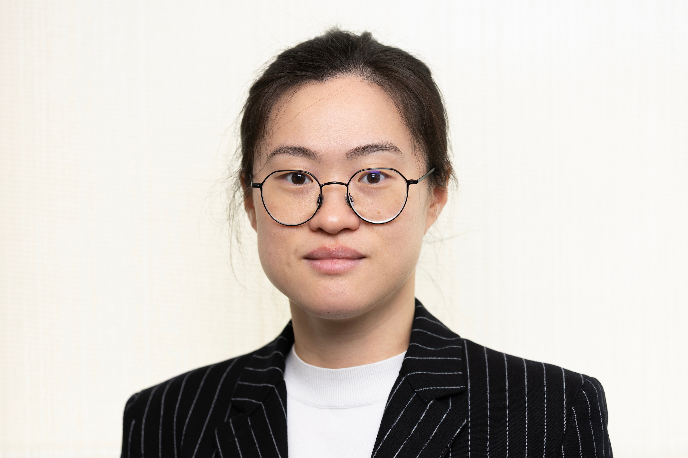 Color portrait of Xiaoqi (Clare) Liu, wearing a black suit jacket with white pinstripes, a white shirt and glasses.