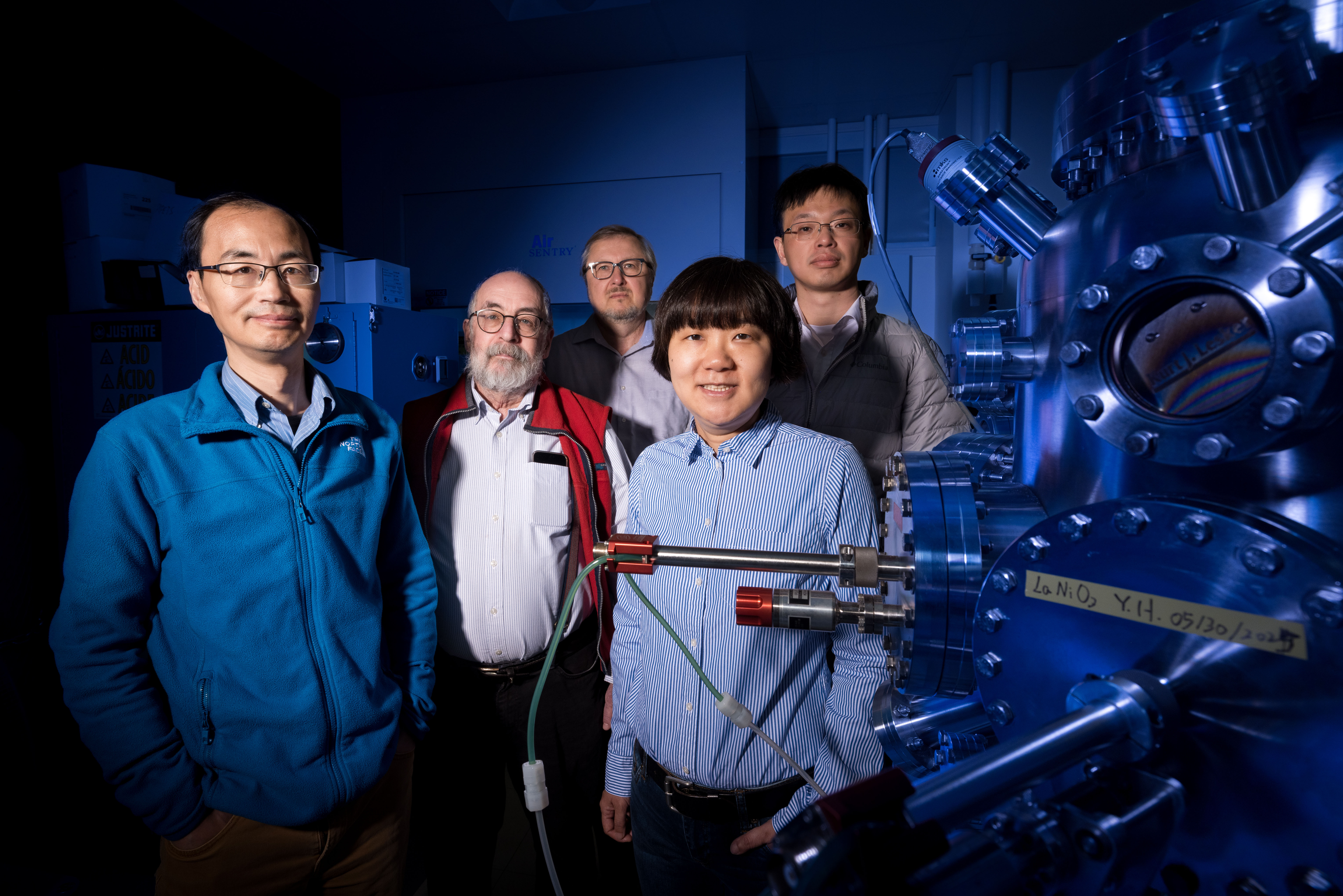 Husker physicists (from left) Xiaoshan Xu, Peter Dowben, Evgeny Tsymbal, Xia Hong and Zuocheng Zhang stand next to an oxide thin film deposition system inside Hong’s lab in Jorgensen Hall. The lighting is blue.