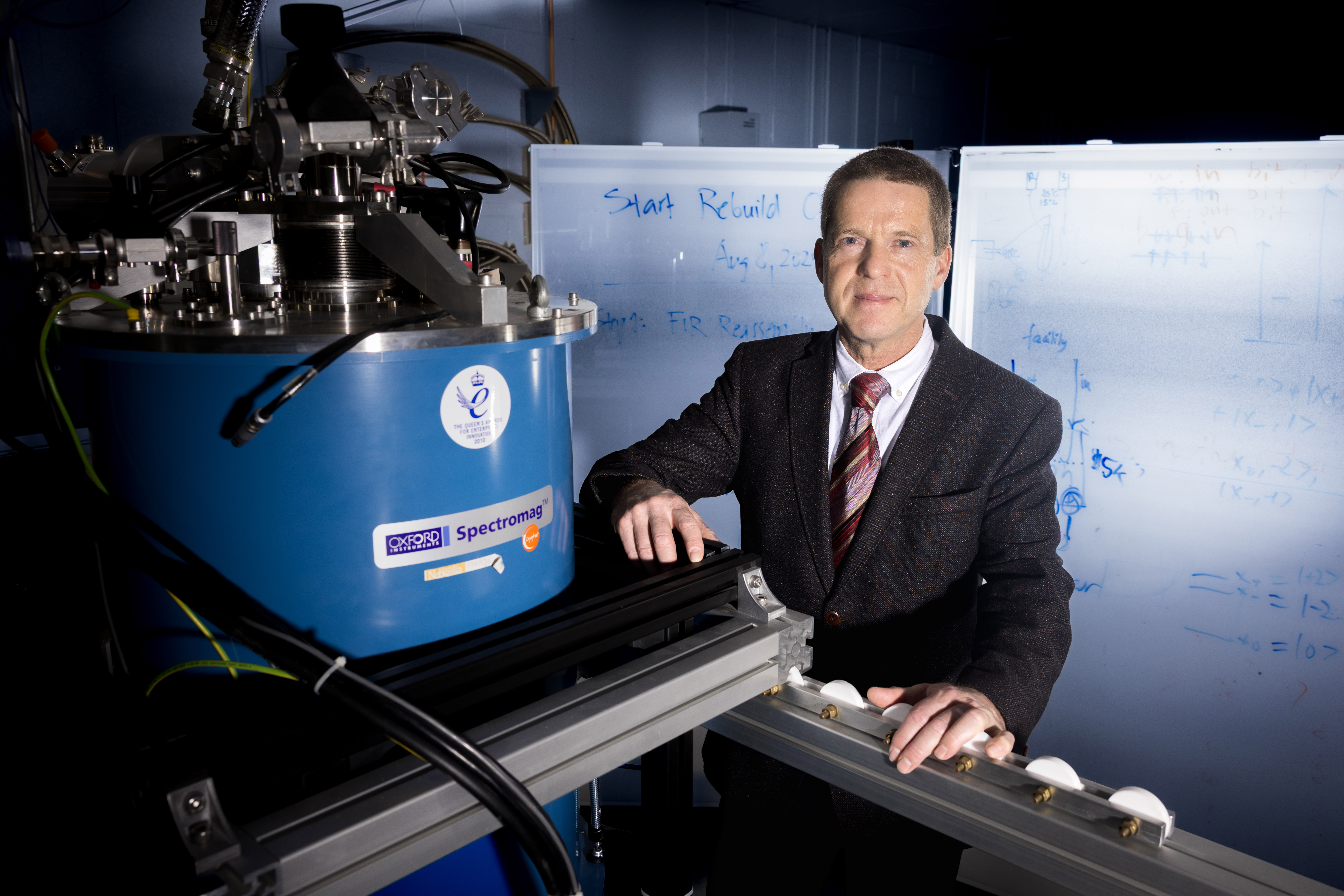 Mathias Schubert, J.A. Woollam Distinguished Professor of Engineering at the University of Nebraska–Lincoln, stands next to an Oxford Instruments Spectromag. He is wearing a dark suit, and behind him are two markerboards with writing on them.