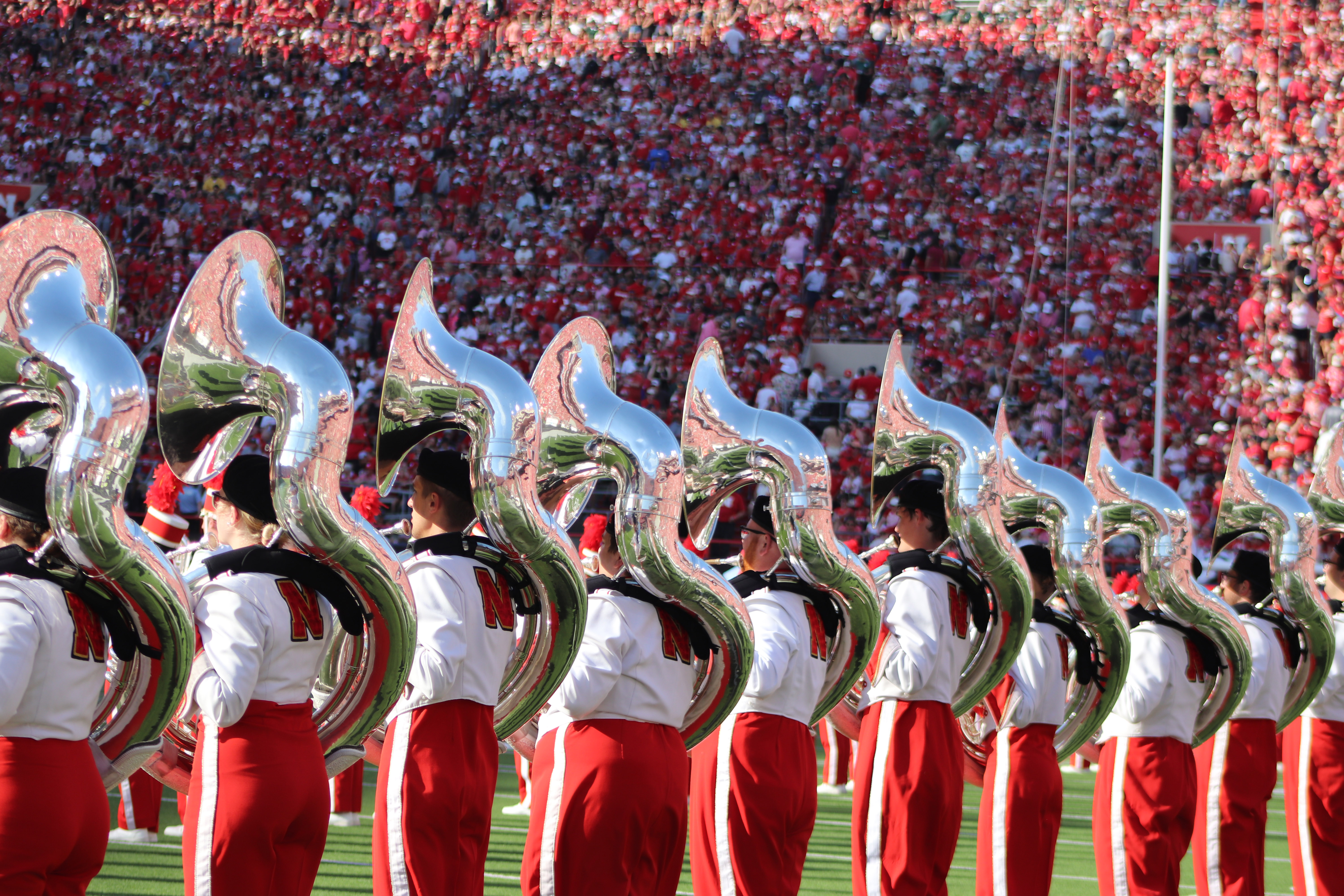 Ten sousaphone players in Cornhusker Marching Band uniforms perform in a line on the field at Memorial Stadium.