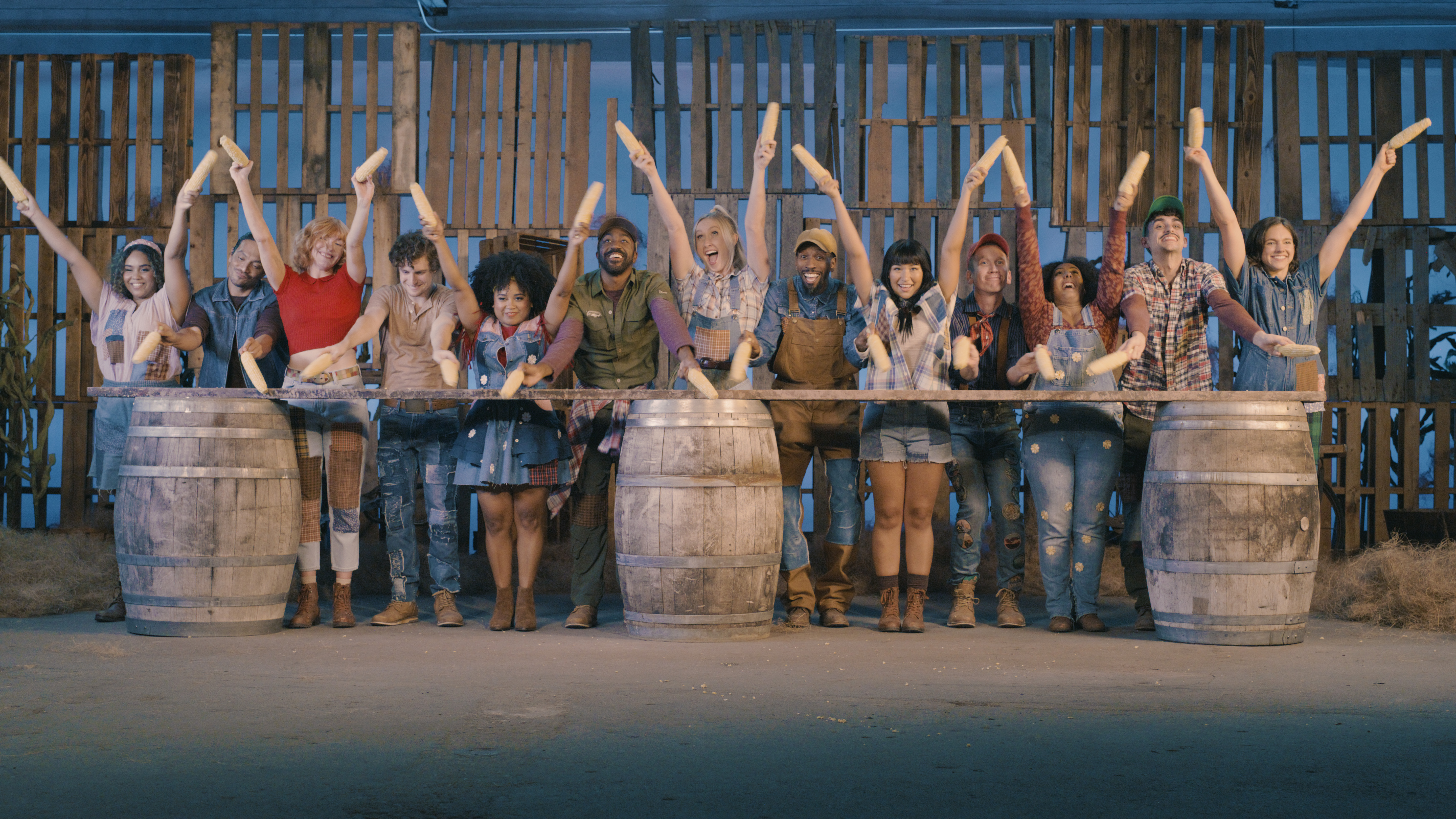 Thirteen actors pose with corn cobs behind three wooden barrels with a board on top in this publicity photo for "Shucked." 