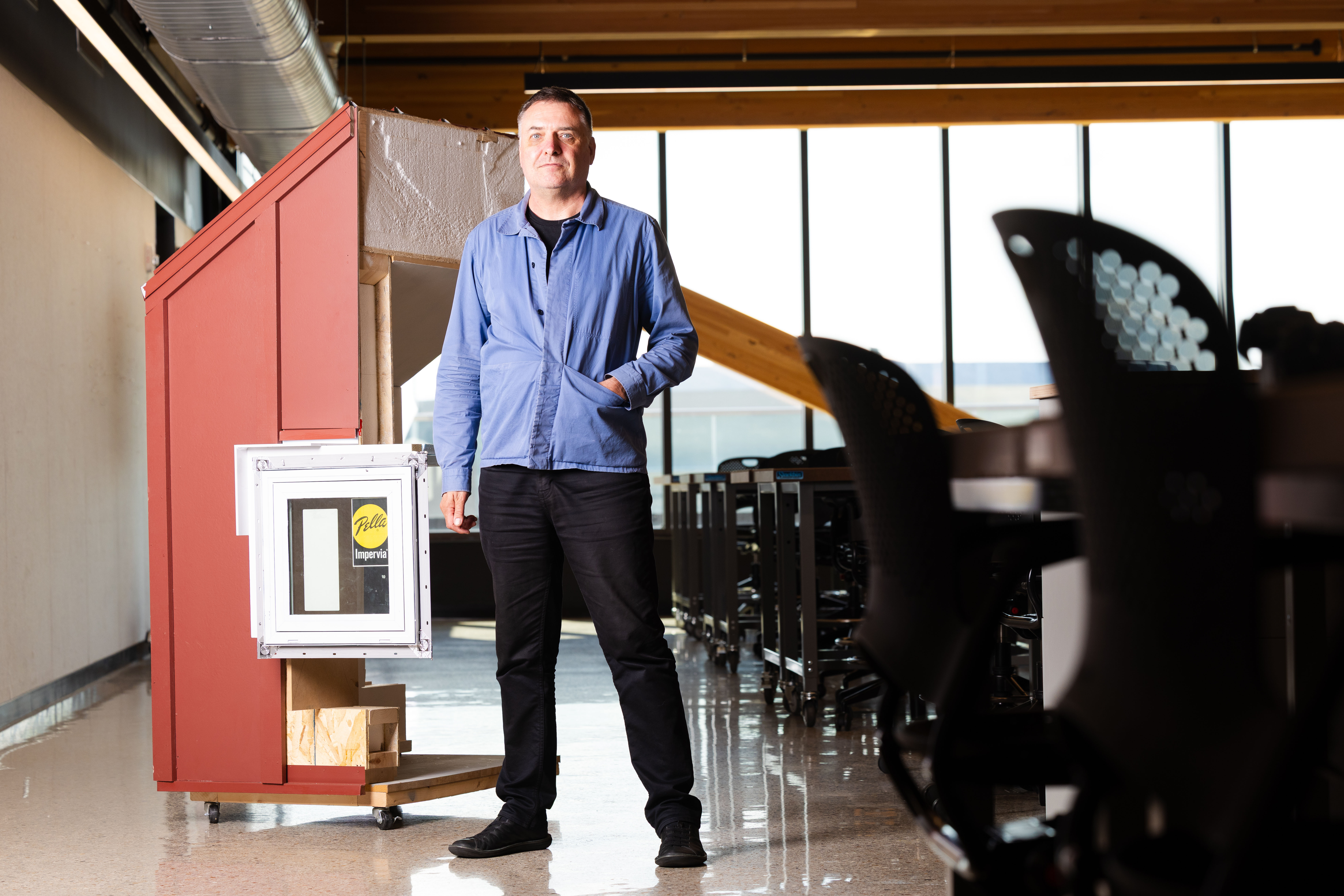 Jeffrey L. Day, professor of architecture at the University of Nebraska–Lincoln, stands in an architecture classroom. He is wearing a blue button-up shirt over a black shirt, black pants and black shoes. His left hand is in his shirt pocket.