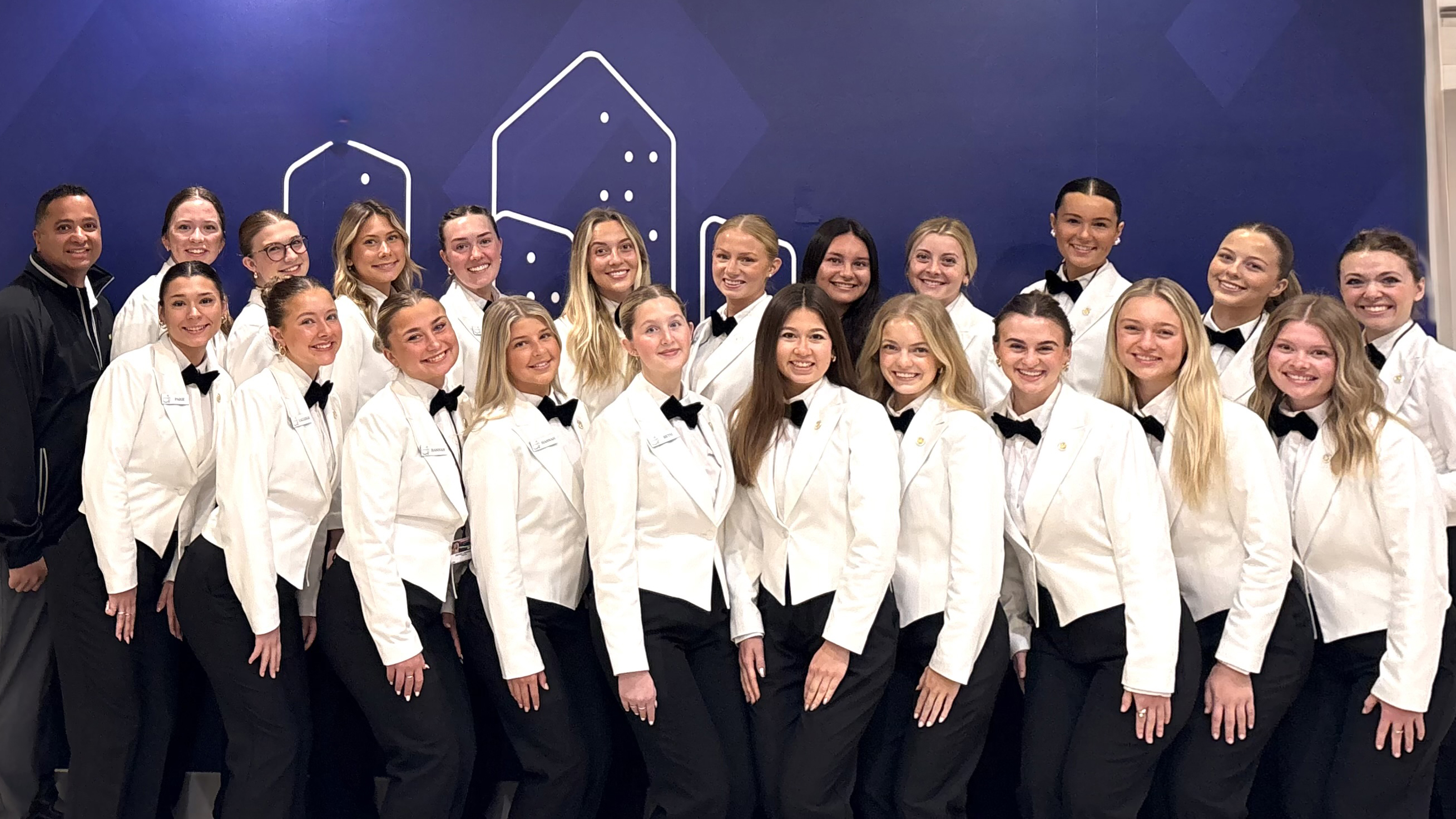 Twenty-one young women wearing white jackets, white shirts, black bowties and black pants pose for a photo with instructor Ajai Ammachathram. They are Husker students providing hospitality services at the 2026 Masters Tournament.