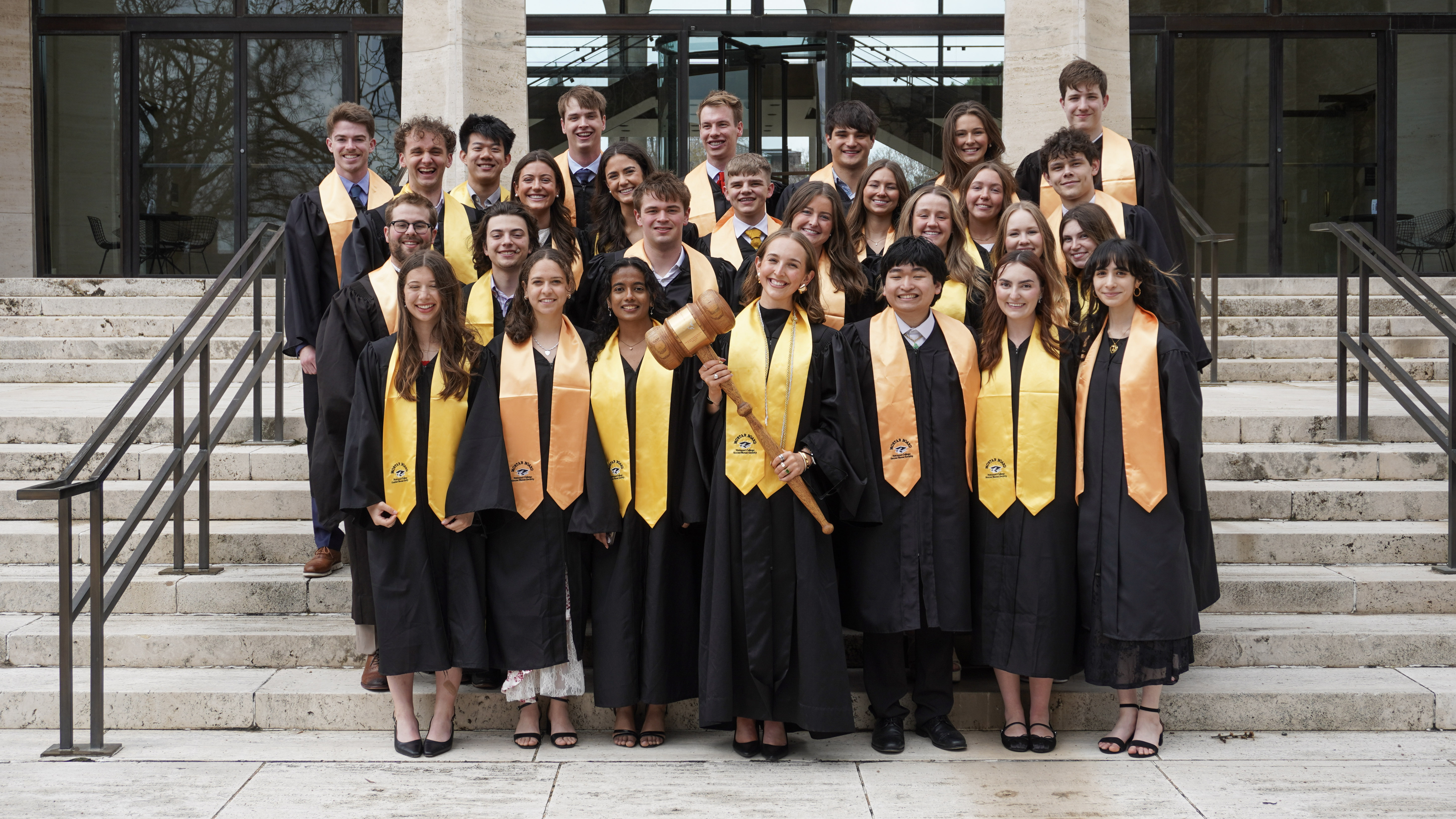 Twenty-eight University of Nebraska–Lincoln students, wearing black robes with gold stoles, pose for a group photo on the front steps of Sheldon Museum of Art. A female student, front-row center, holds an oversized gavel.