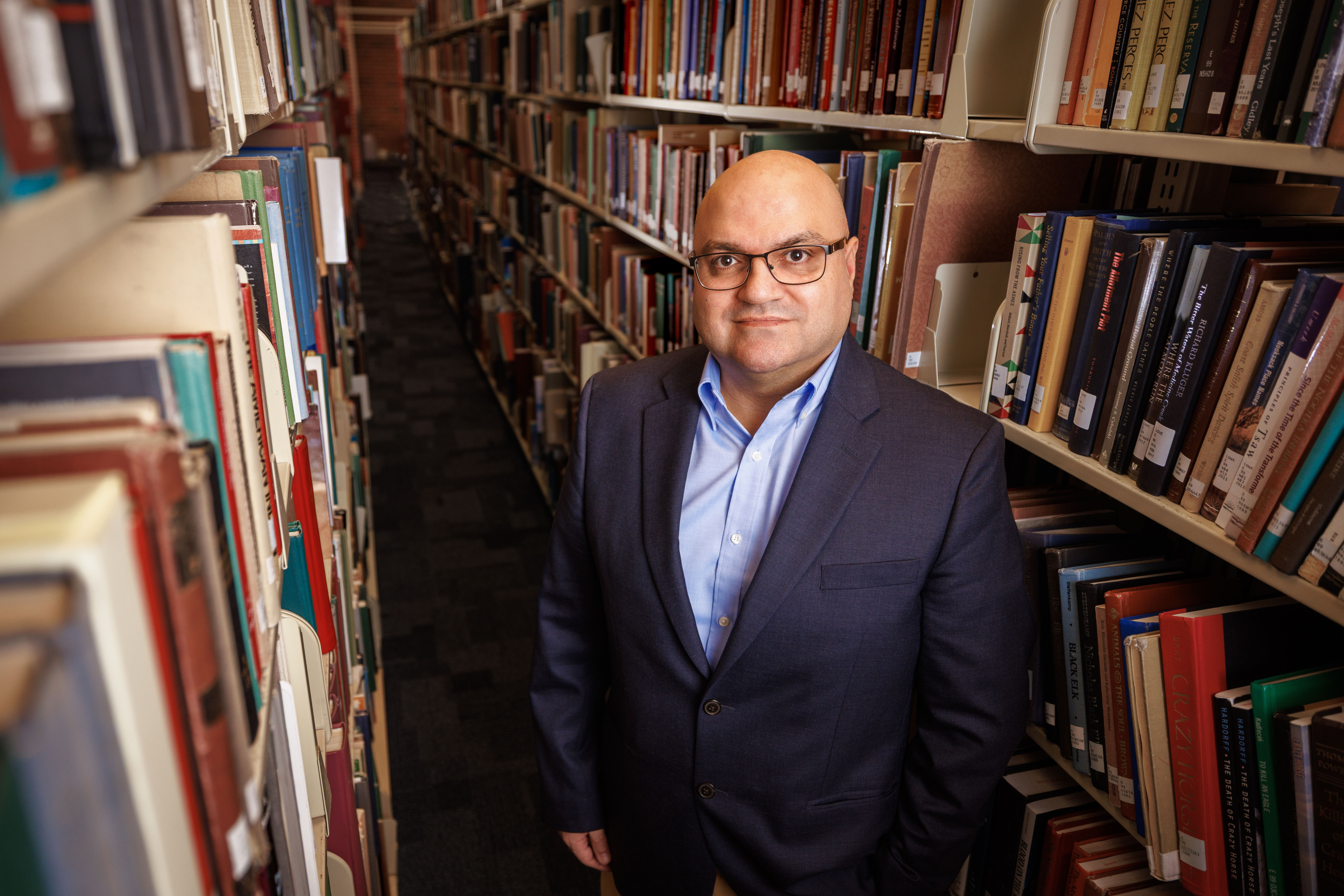 Bedross Der Matossian, Hymen Rosen Professor in Judaic Studies at the University of Nebraska–Lincoln, stands between rows of bookshelves in Love Library. He is wearing a dark blue sport coat over a light blue dress shirt and glasses.