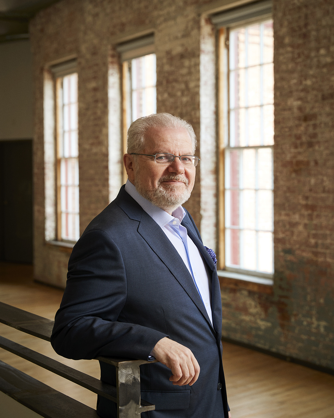 Color portrait of pianist Emanuel Ax, leaning against a wooden rail in a room with brick walls and three tall windows. He is wearing a blue suit coat with a light-colored dress shirt and glasses.