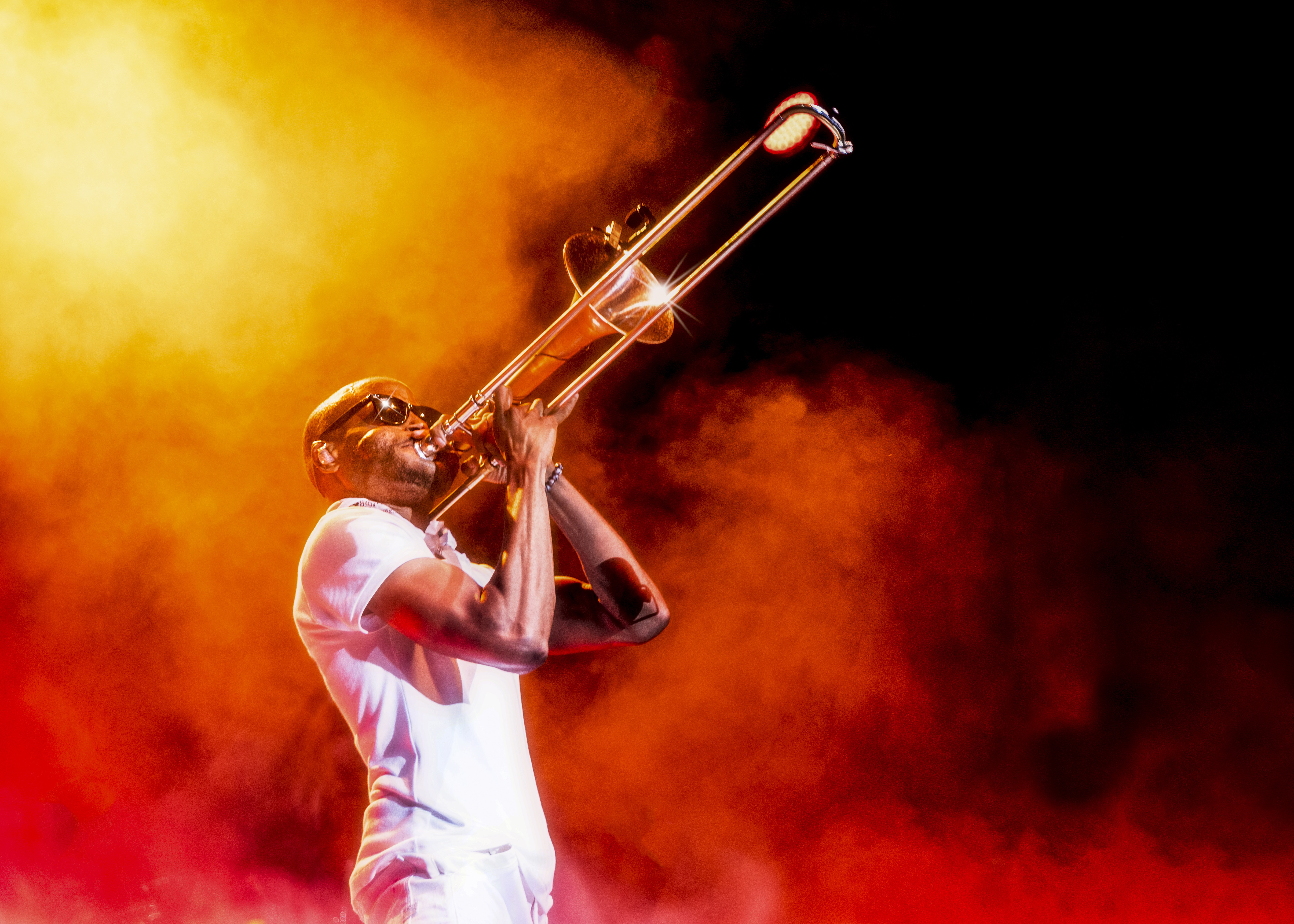 Musician Trombone Shorty, wearing a white short-sleeve shirt and white pants, plays a trombone on a smoke-filled stage.