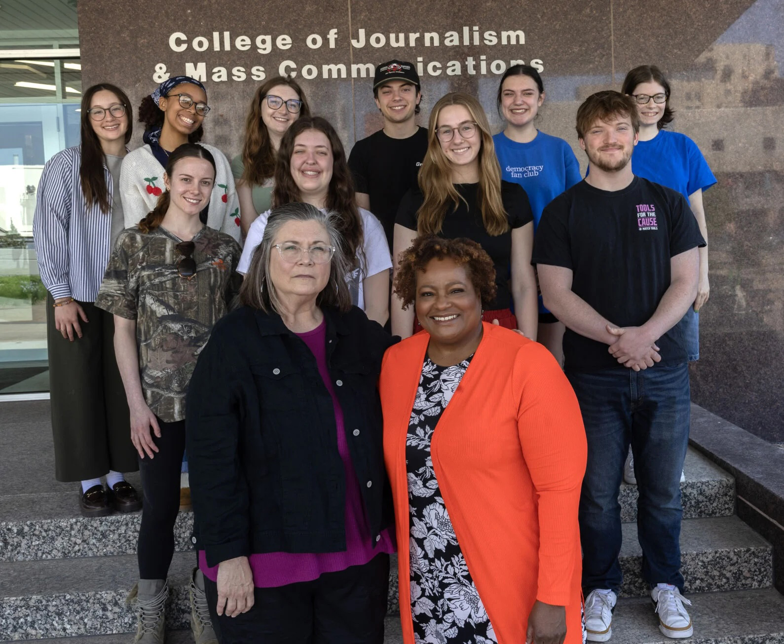 Ten Husker students — eight women and two men — and two female instructors pose for a group photo in front of Andersen Hall. The words "College of Journalism and Mass Communications" can be seen on the exterior wall.