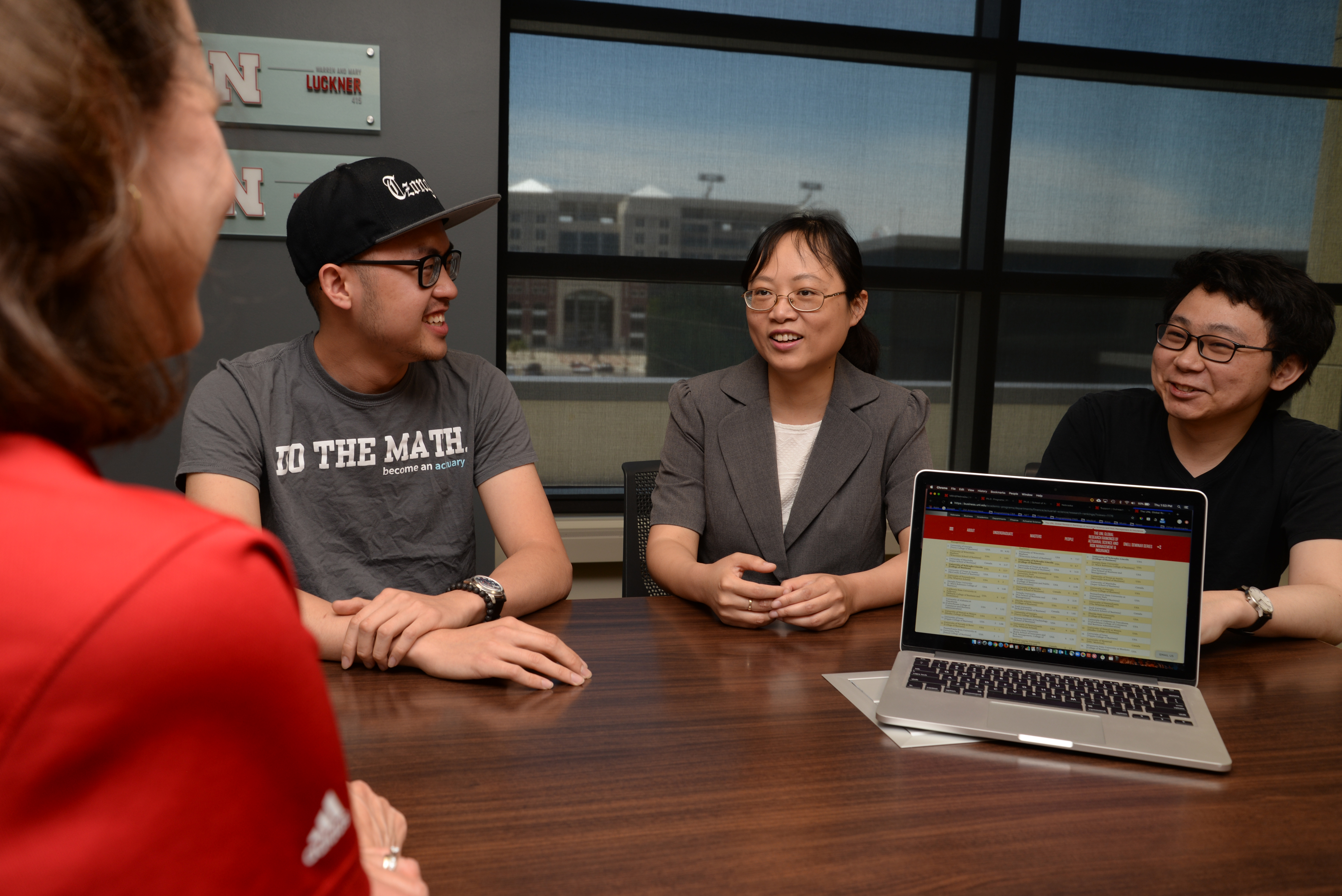Two men and two women sit around a table, with an open laptop on it. Memorial Stadium can be seen through a window in the background.