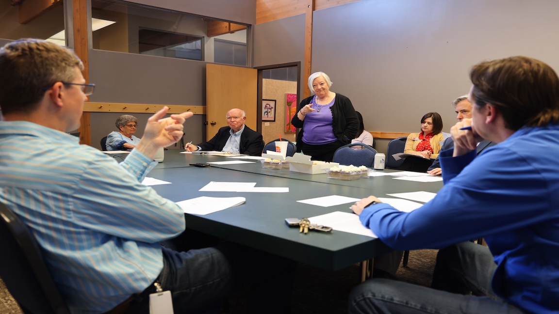 Mary Emery stands in a purple shirt and black jacket around a crowded table of men and women.