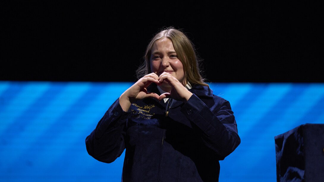 Claire Woeppel, an ALEC sophomore from Chambers, dressed in her dark blue corduroy FFA jacket, forms a heart with her hands. 