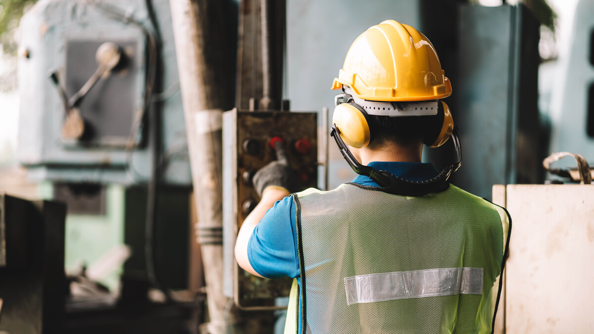 A man wearing a vest, hard hat and ear protection presses a button on a control panel.