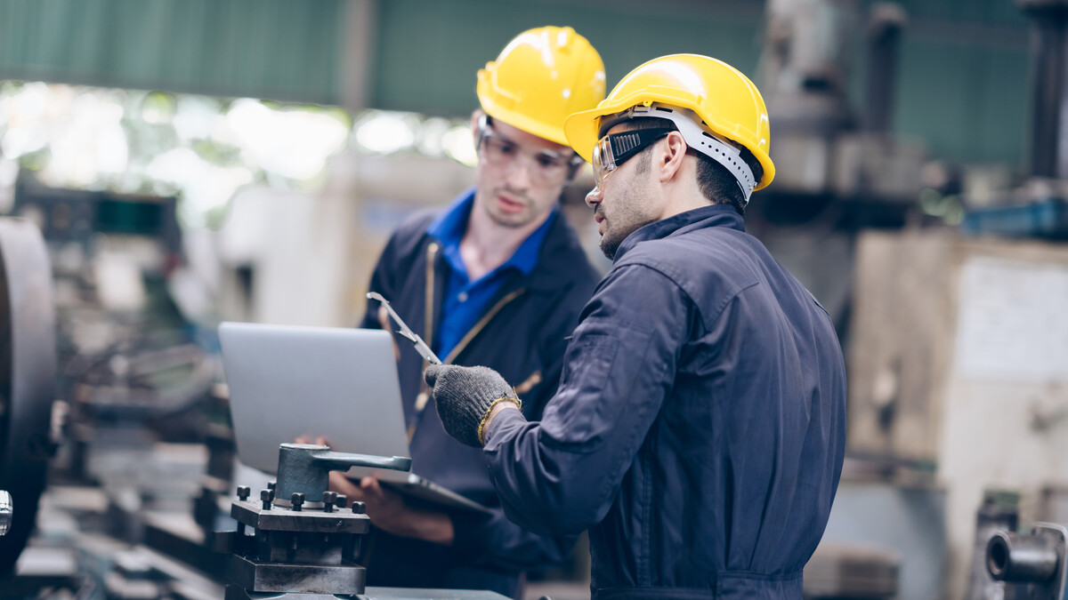 Two male manufacturing workers in dark blue jumpsuits, yellow hard hats and safety glasses confer.