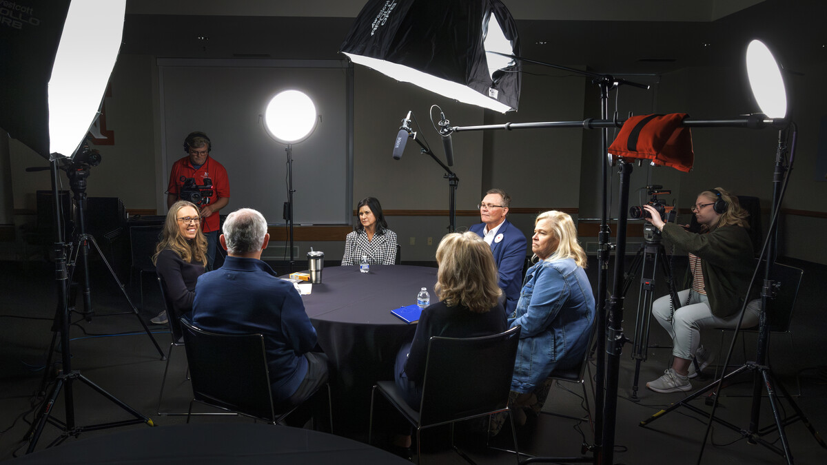 Four women and two men sit at a round table in a dark room, surrounded by lighting umbrellas, as a man and woman film.