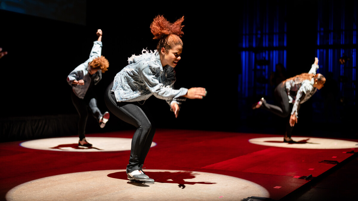 Three young women tap dance on a dark stage, illuminated by three spotlights.