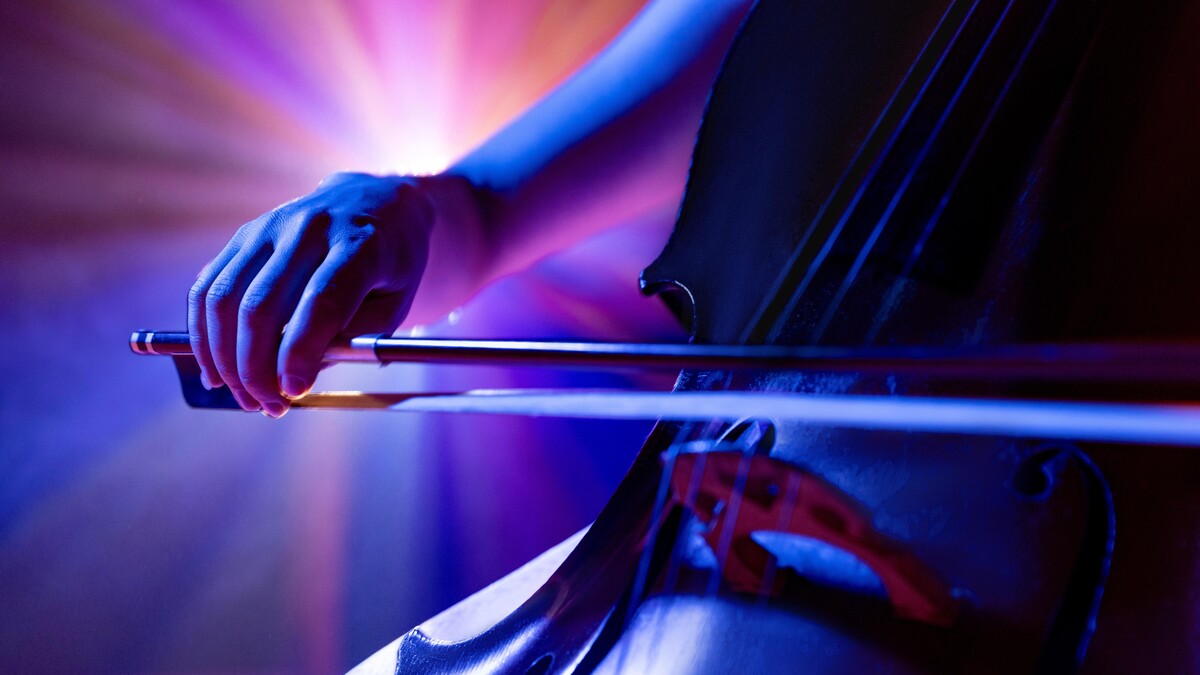 Close-up of a hand pulling a bow across a cello's strings, with a spotlight in the background.