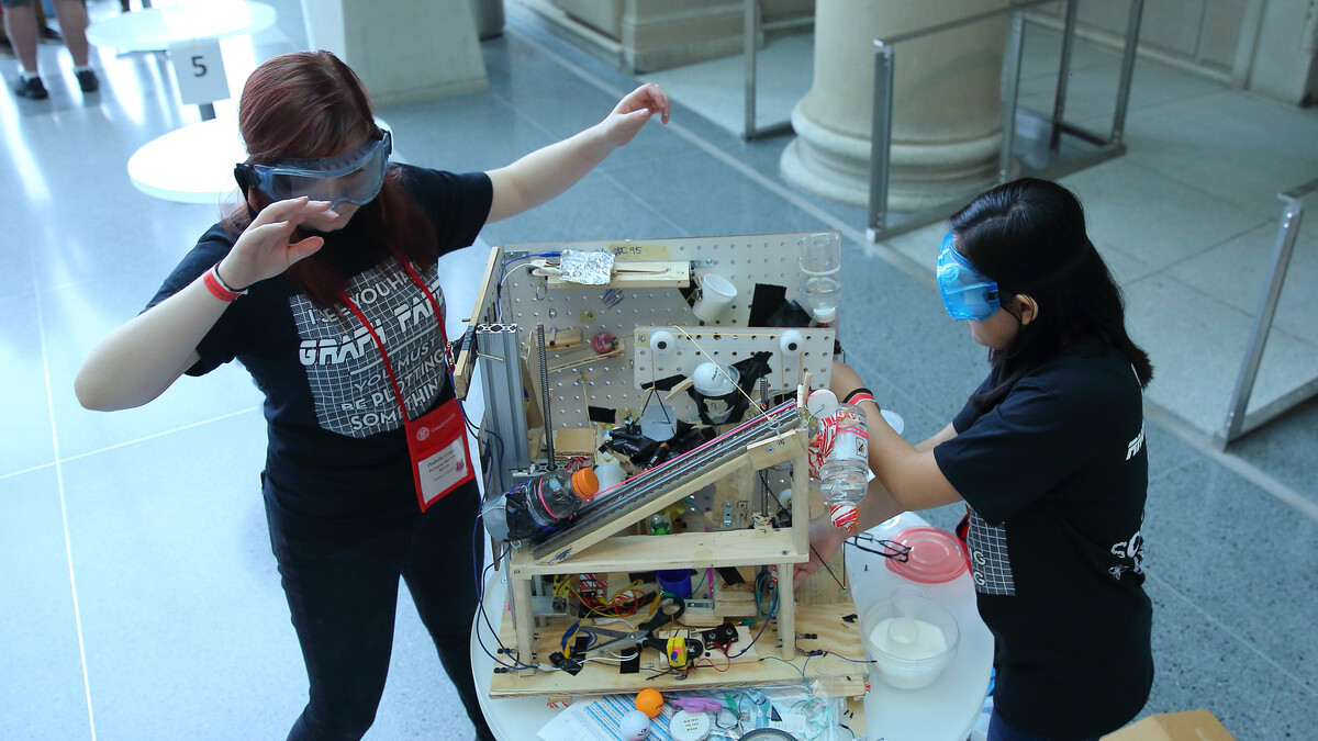 Two girls dressed in black, wearing safety glasses, adjust a Rube Goldberg-style device.
