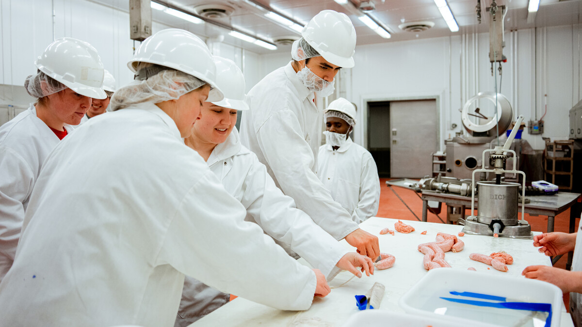 Students in the UNL Meat Science Club — wearing labcoats, hair nets and white hard hats — work with a sausage maker. Several sausages lie on the table.