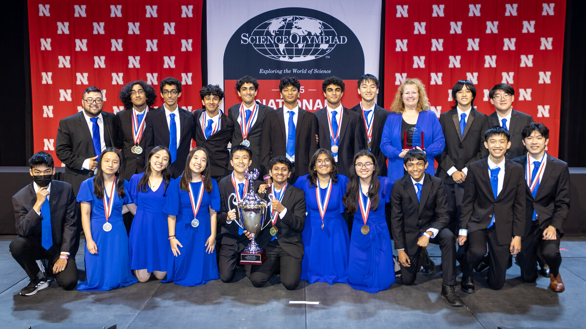 Twenty students and their coaches from Seven Lakes High School in Katy, Texas, wearing formal attire, pose with a large trophy in front of a Husker background and the Science Olympiad National Tournament logo.