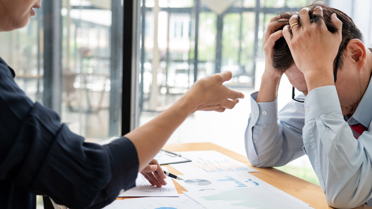 A woman gestures toward a distressed man with his head down and his hands on top of his head in a workplace.