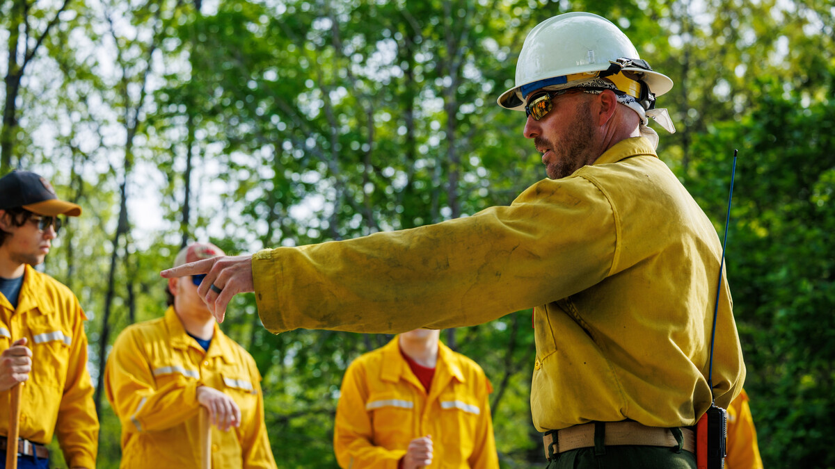 Eric Moul, fire management specialist with the Nebraska Forest Service, points. He is wearing a yellow shirt and white hard hat. Three other people stand in the background, also wearing yellow shirts.
