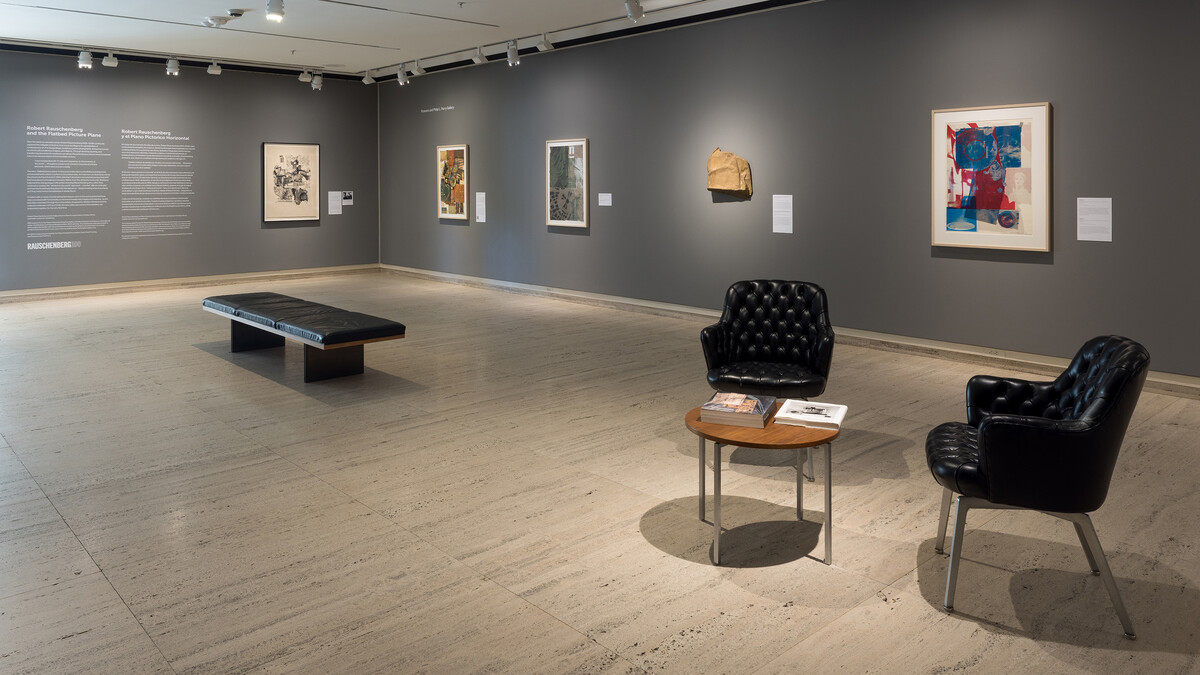 An art gallery at Sheldon Museum of Art with a bench, two chairs and a small table. There is text on the far wall and five artworks visible on the adjoining wall.