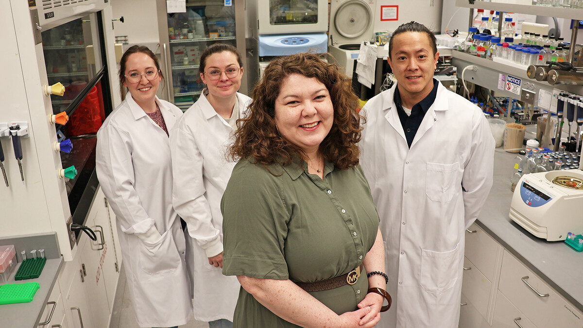 Four University of Nebraska–Lincoln researchers — (from left) Darla Brennan and Madison Broeker, doctoral students in biochemistry; Nicole Buan, professor of biochemistry; and Henry Chen, postdoctoral researcher in biochemistry — pose in a lab. Buan is wearing a green dress and the others are wearing white labcoats.