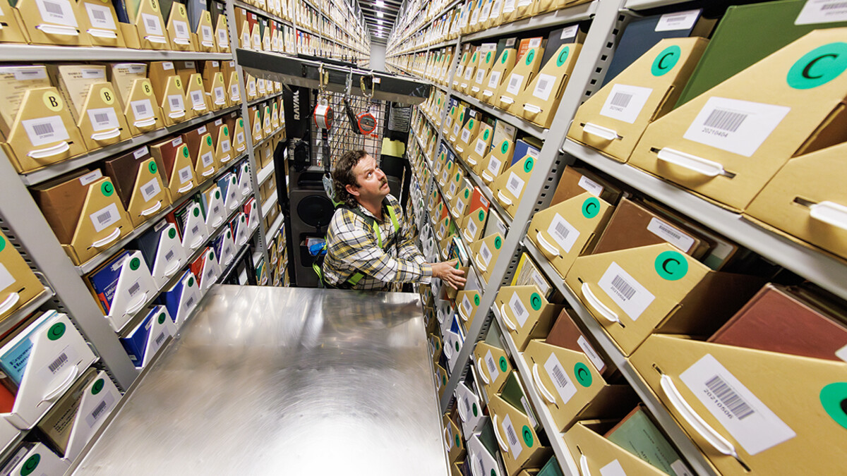 A man stands on a cherry picker between large shelves of documents.