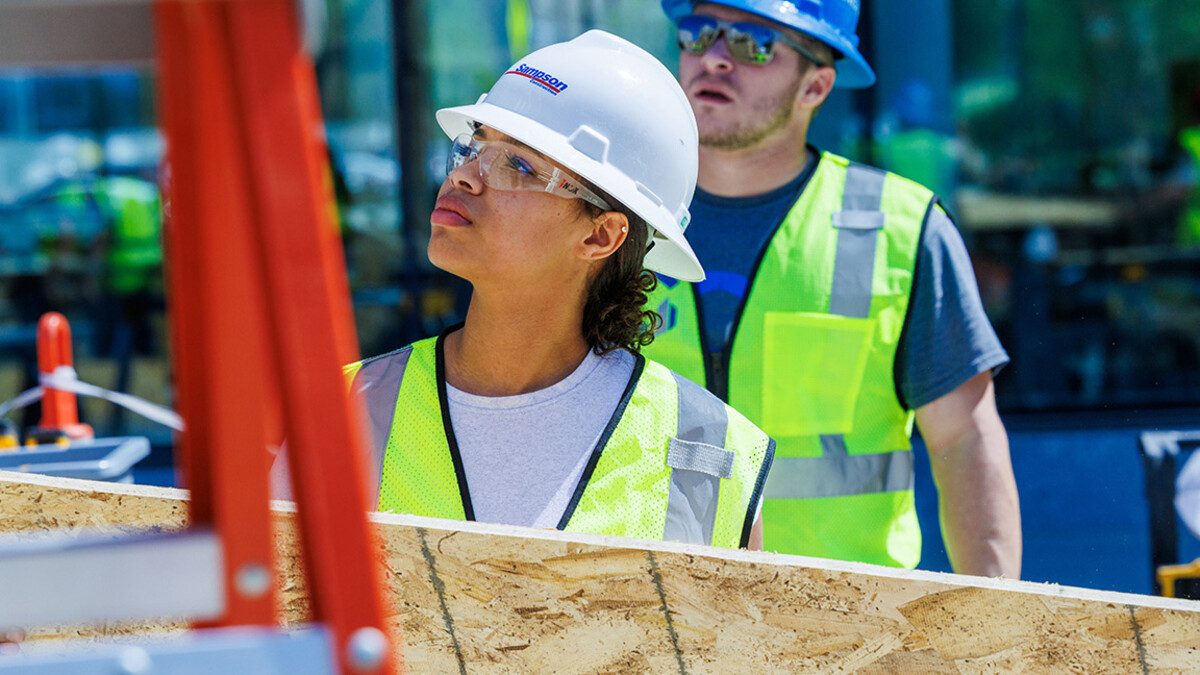 A black woman and a white man in t-shirts, construction vests, white hard hats and clear protective glasses stand on the left of a photo next to a ladder.