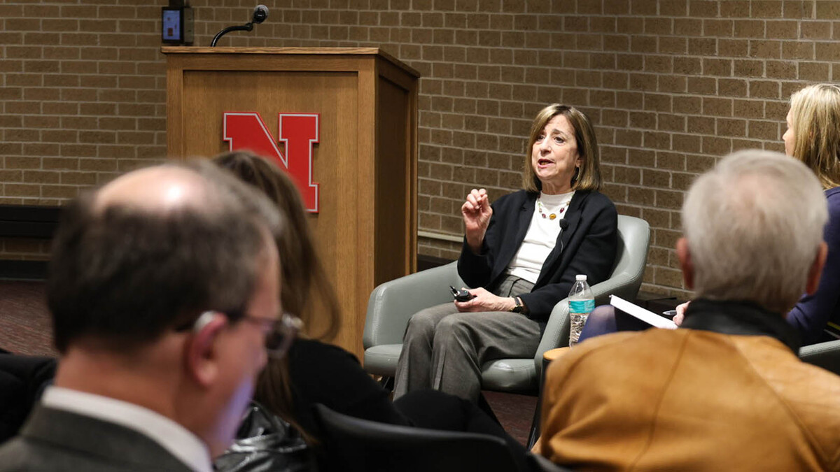 A white, brunette woman in brown slacks, a white shirt and a black blazer sits, speaking in front of a crowd.