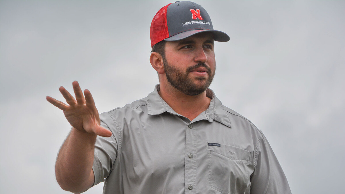 Jacob Harvey, a white man with a brown beard, gestures while out in a field under a gray sky, wearing a gray shirt and gray and red Nebraska trucker hat.