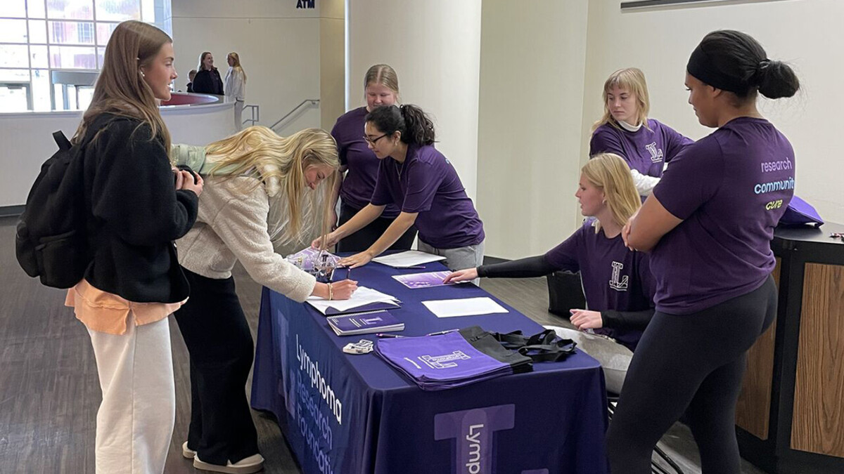 CoJMC students in purple shirts gather behind a booth, speaking to two students across the table.