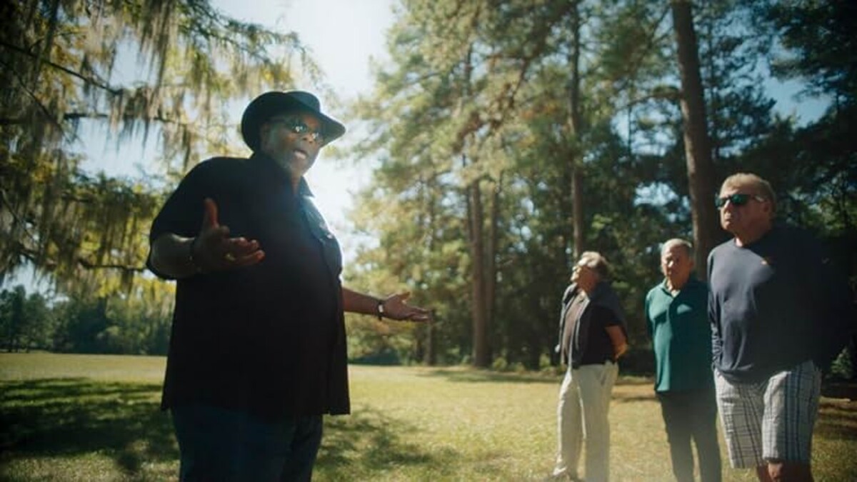 A tour guide, a Black man in a black shirt and black cowboy hat, stands in front of a small crowd outside surrounded by tall trees.