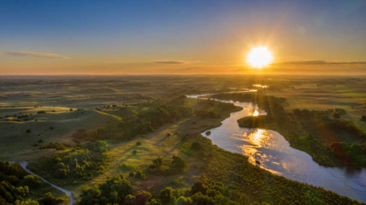 Green flat land with a river and shrubbery and trees and a sunset in the background
